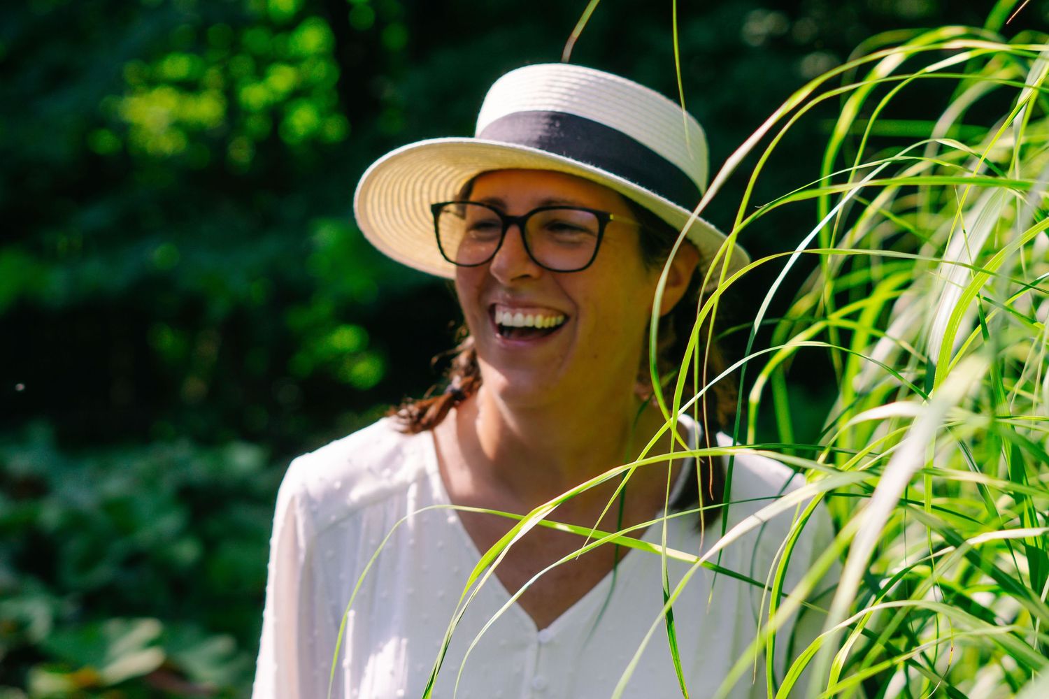 A joyful expression shines through glasses and a sun hat against a lush green garden background.