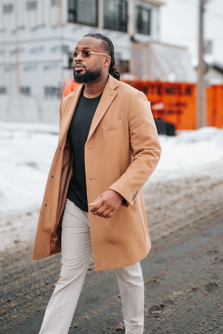 Stylishly dressed person in camel coat and white pants walking on snowy street with orange construction barrier visible.