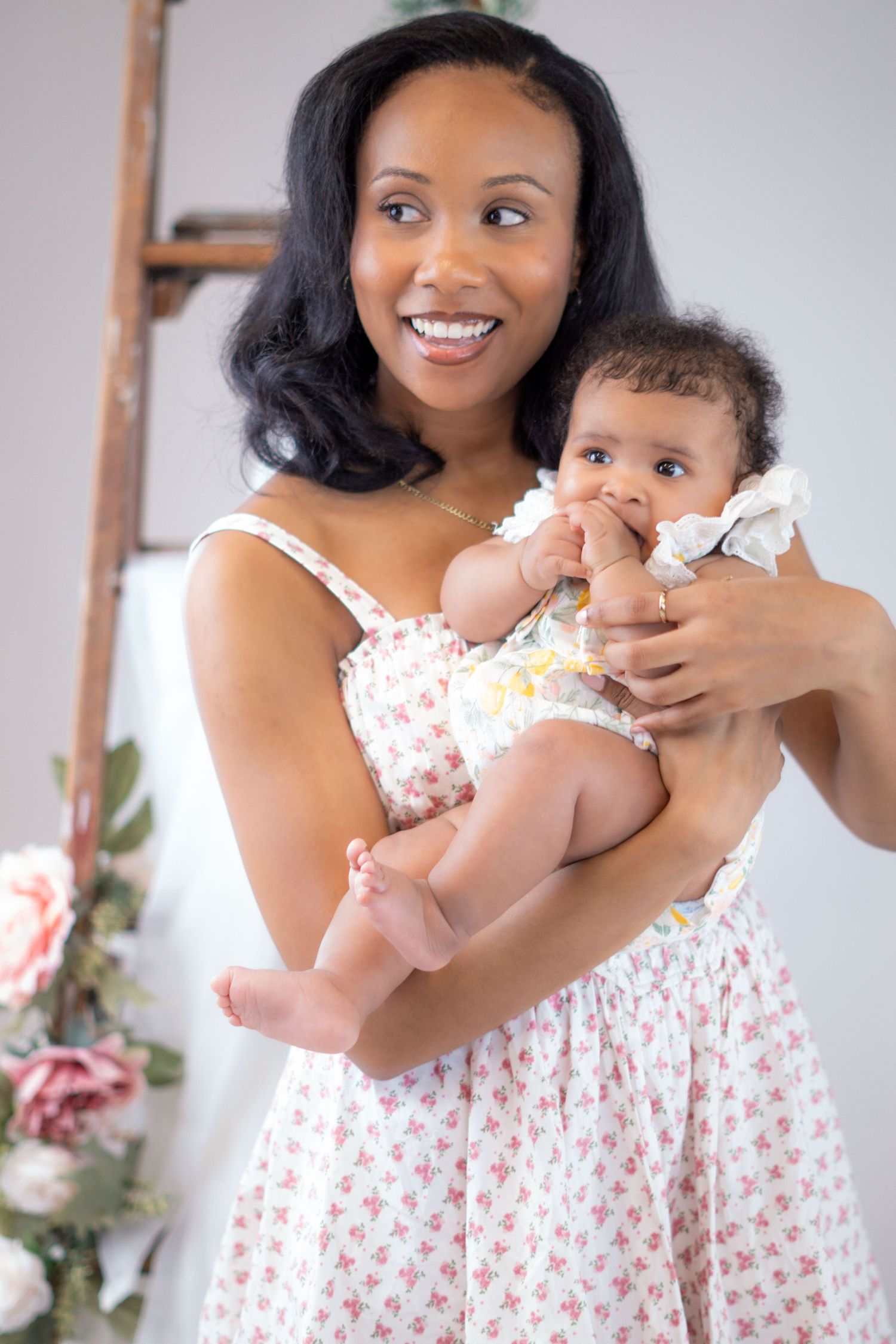 A mother in a floral dress holds her baby who wears white, standing in front of a decorative floral background.