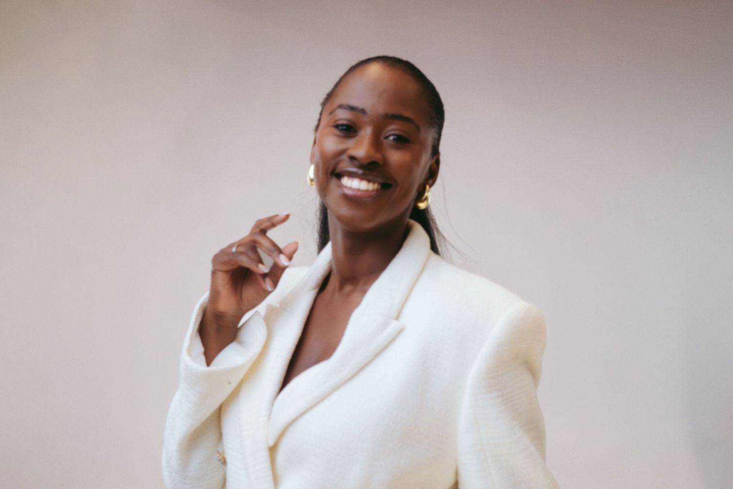 Professional wearing a white blazer smiles warmly against a neutral background in a studio photoshoot.