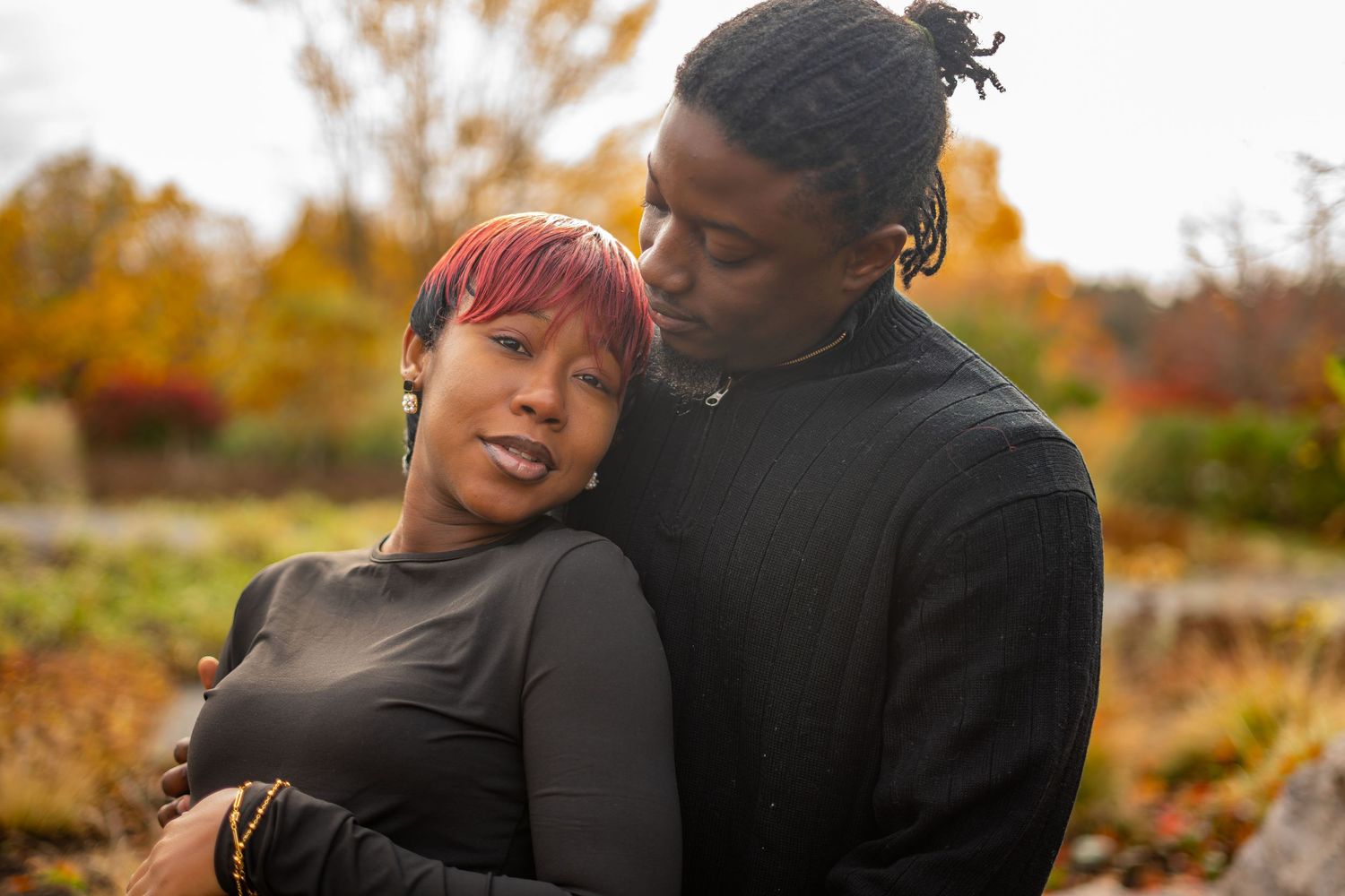 A couple embraces outdoors during autumn with orange and yellow fall foliage in the background.