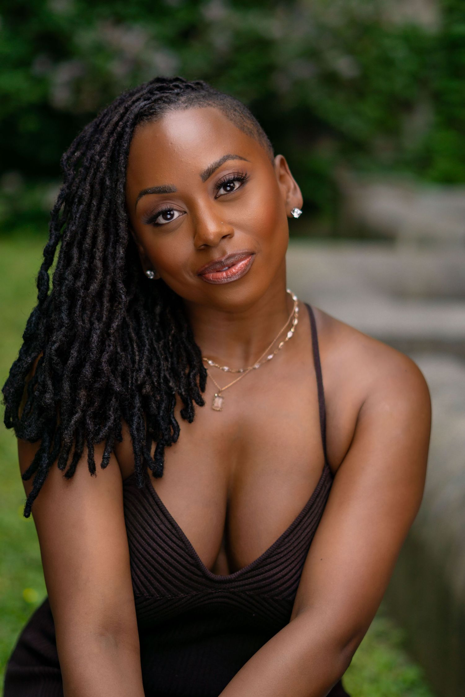 Portrait outdoors featuring long locs hairstyle and black sleeveless top against natural greenery background.