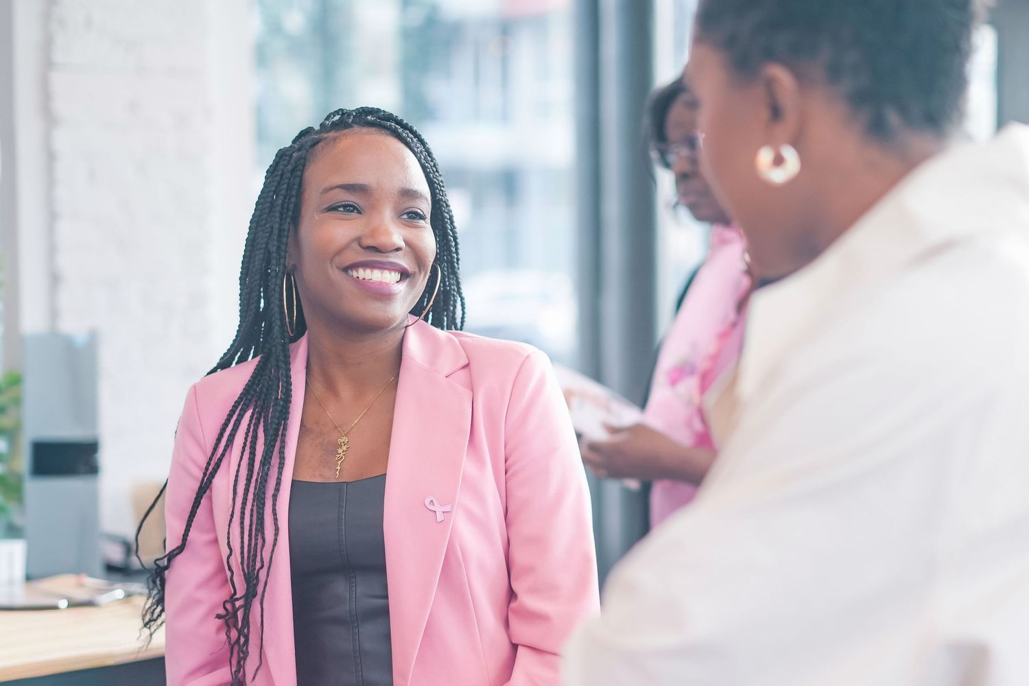 Business professionals in pink blazers engage in animated conversation during a meeting at a modern office.