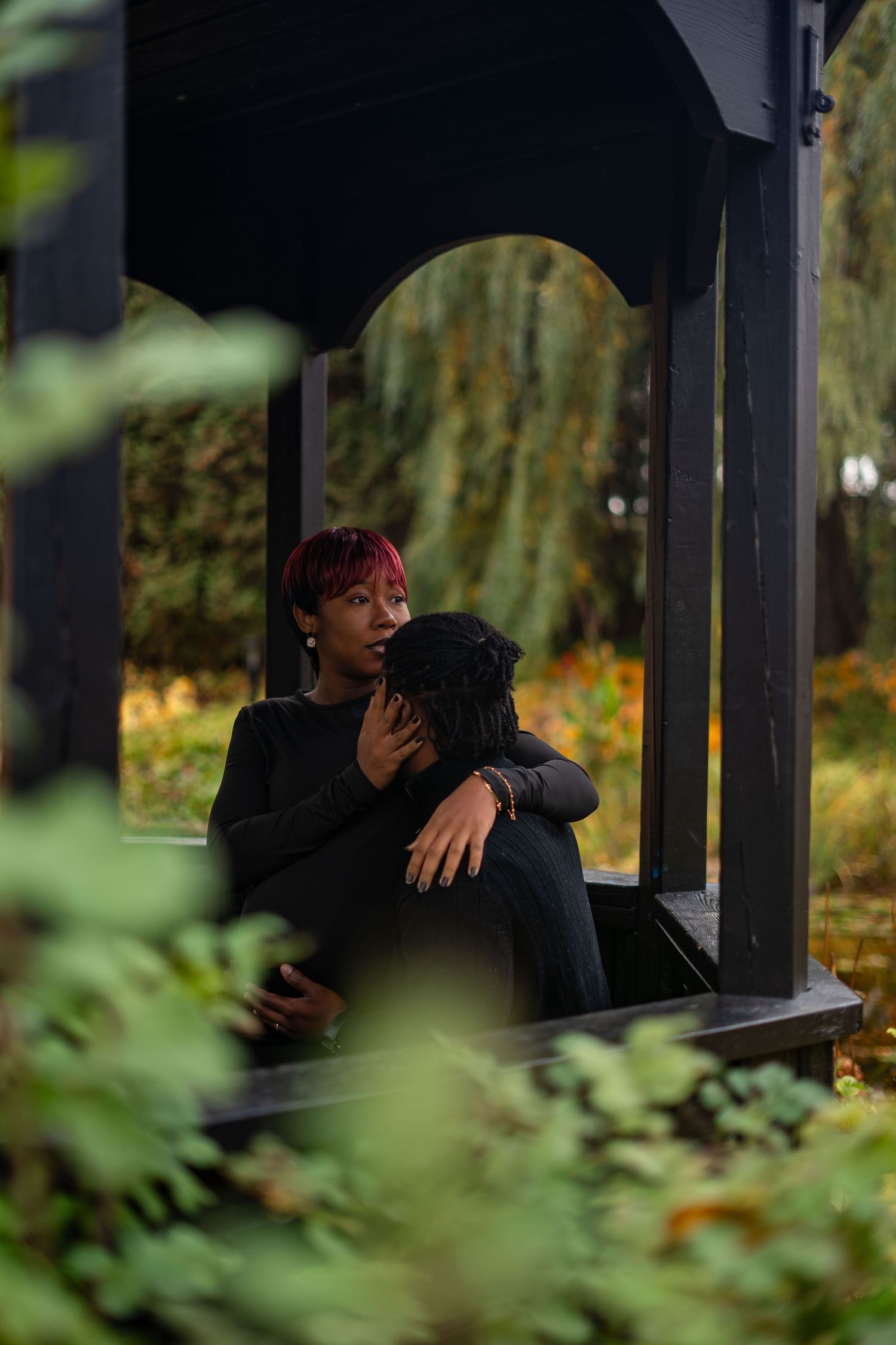 A person sits alone in contemplation on a wooden bench surrounded by lush greenery and dark shadows.