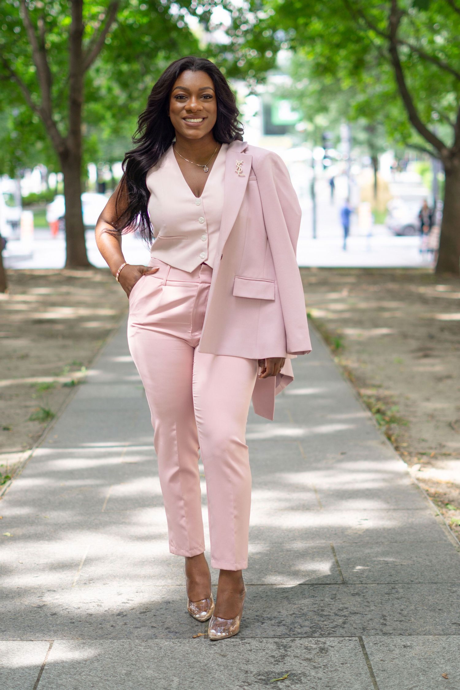 Person in a light pink business suit walks confidently along a tree-lined pathway.