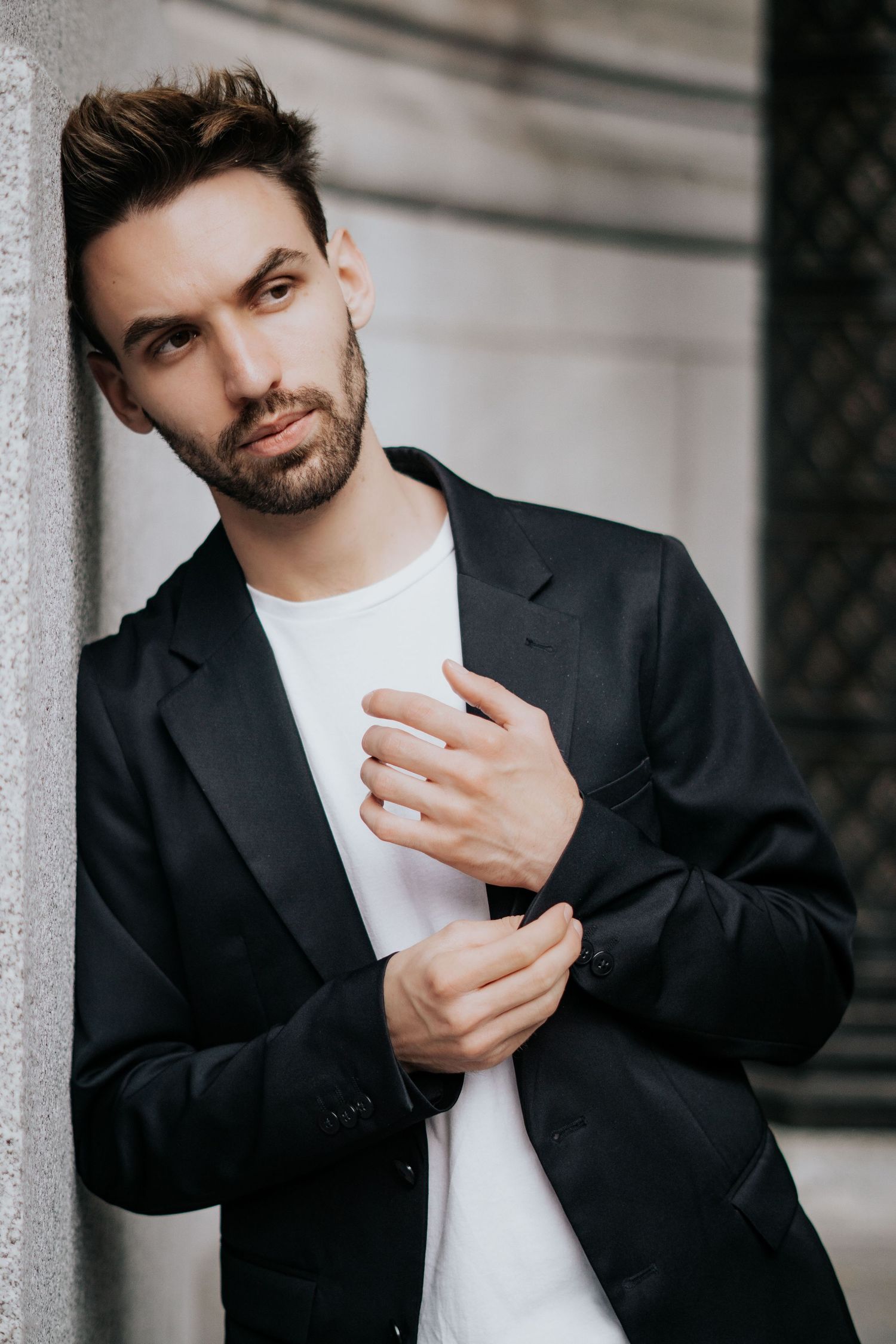 Stylish portrait of person in black blazer and white t-shirt adjusting sleeve cuff against grey wall.