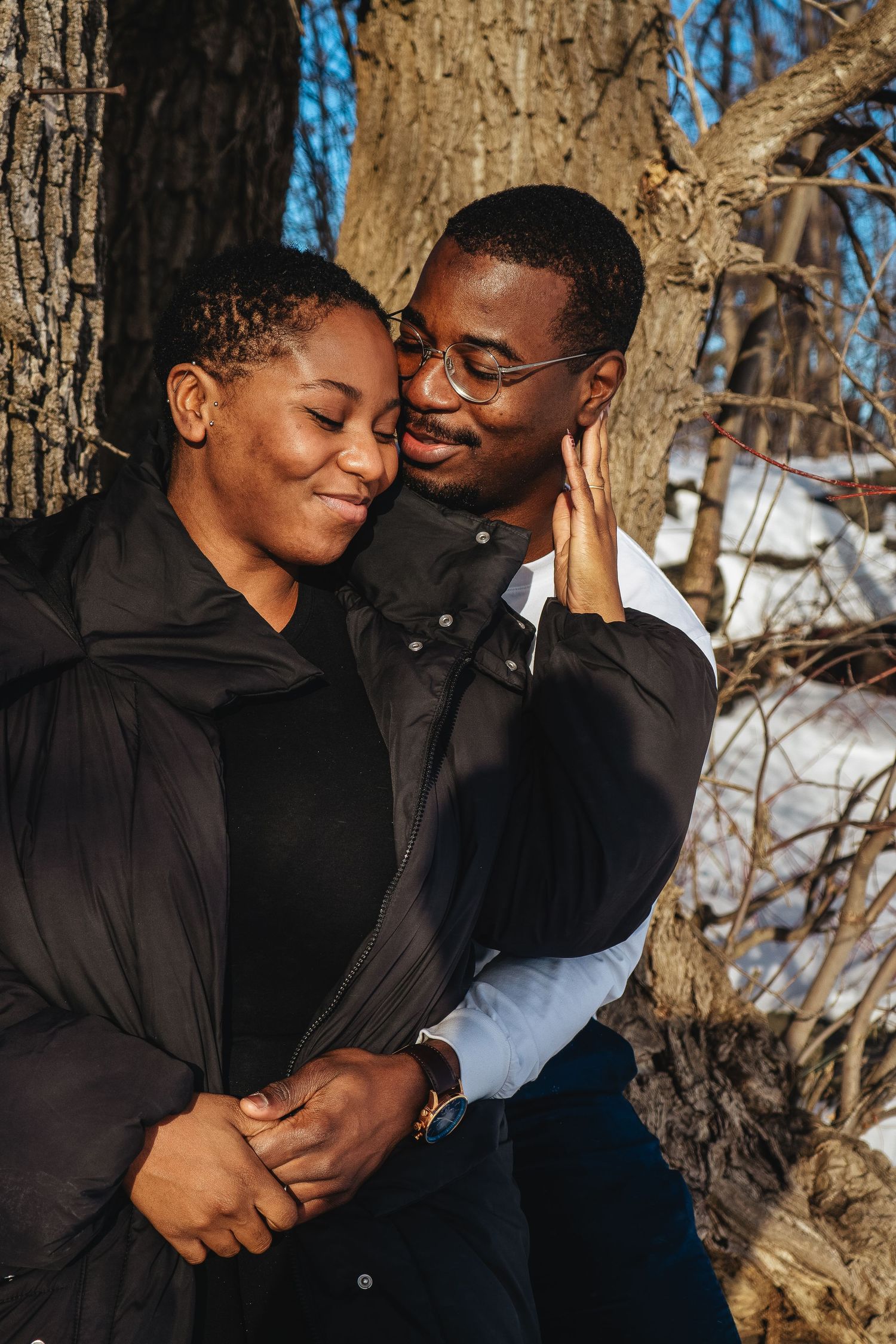 A couple shares an intimate moment outdoors against a tree trunk in winter.