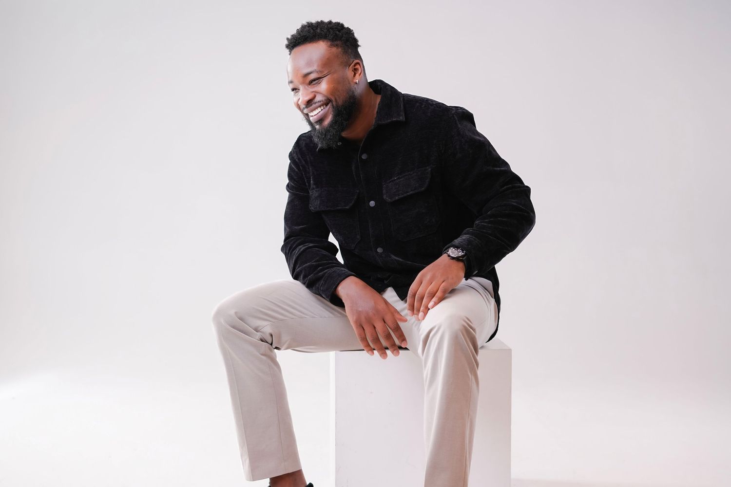 Someone wearing a black sweater and light colored pants sits casually on a white chair against a light background.
