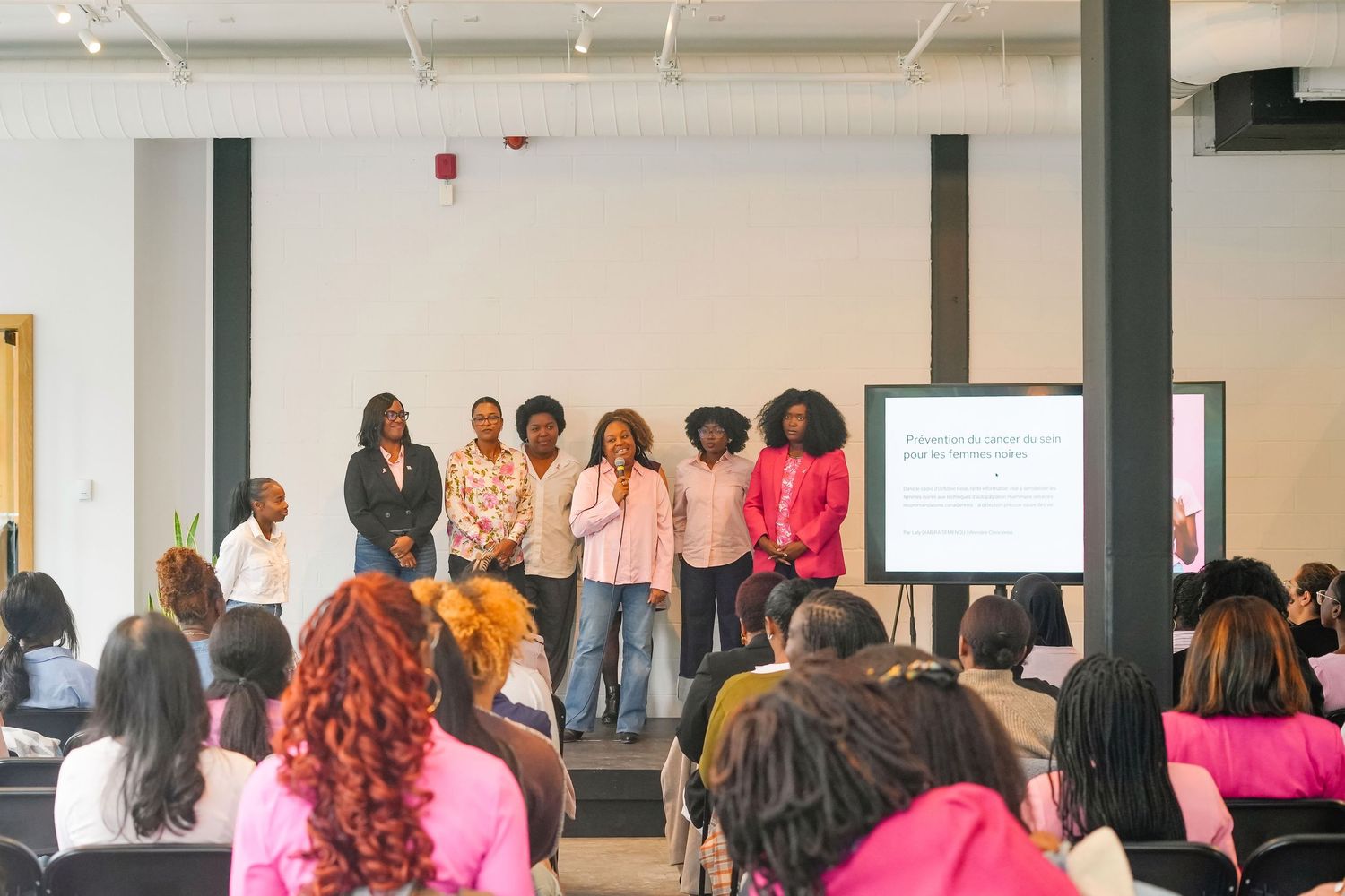 People gather in a conference room for a professional event with pink-clad attendees and presentation screens visible.