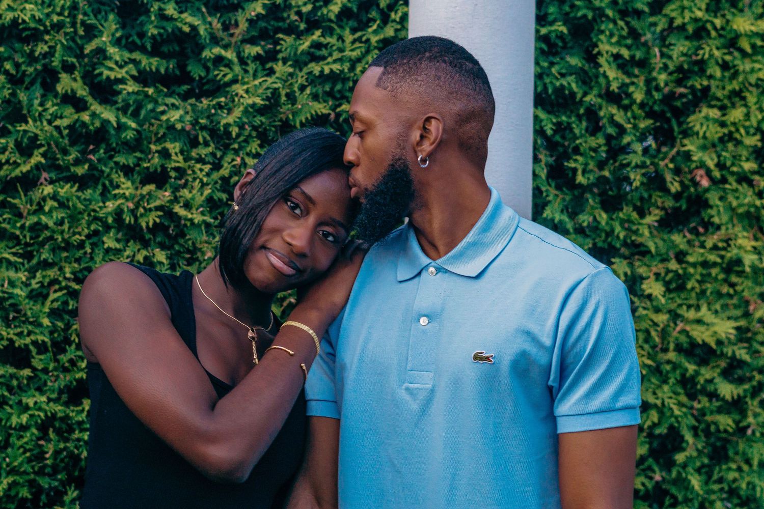 A couple shares an intimate moment in front of a lush green hedge while wearing coordinated blue outfits.