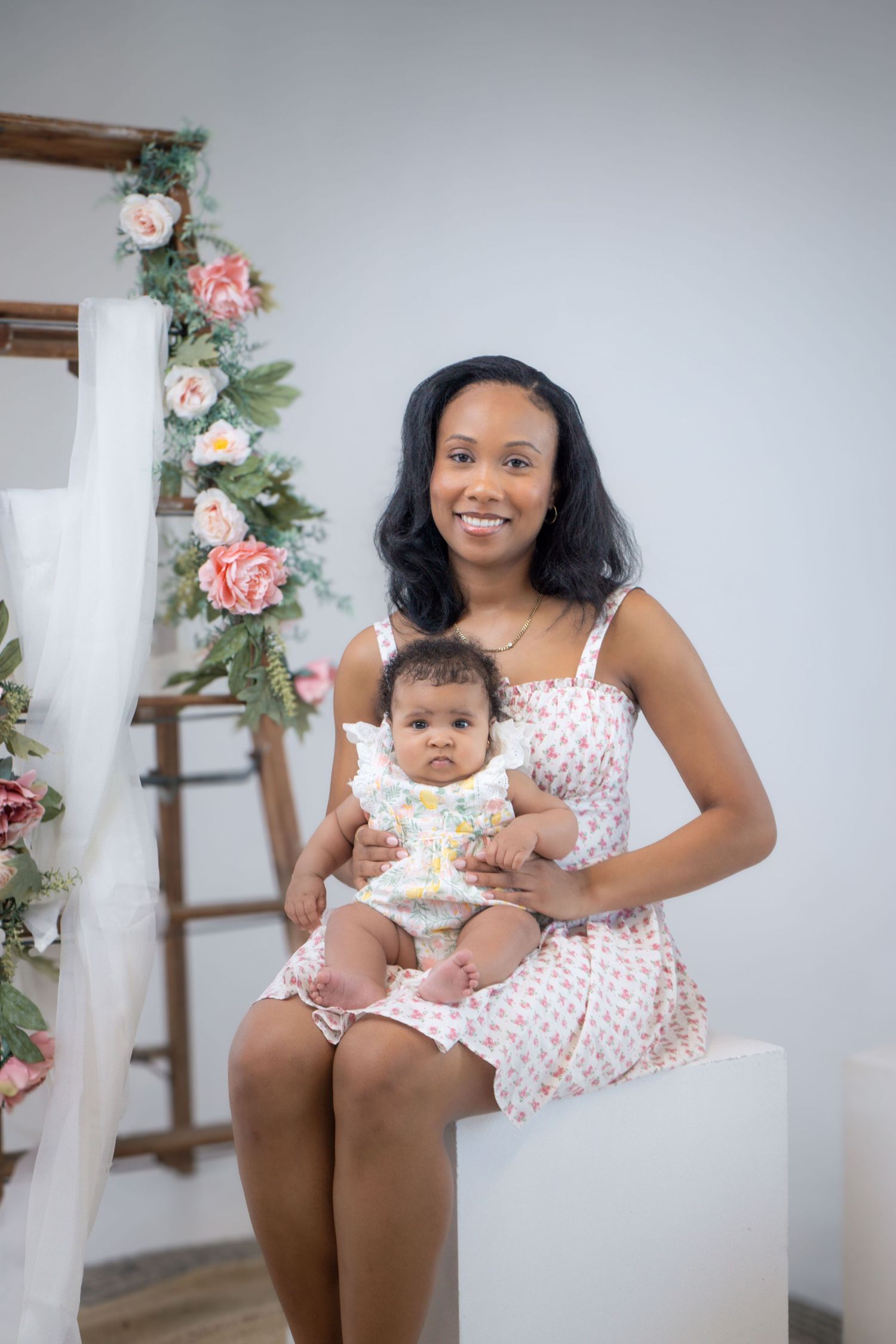 Parent sits with baby on white pedestal near wooden ladder with pink floral garland.