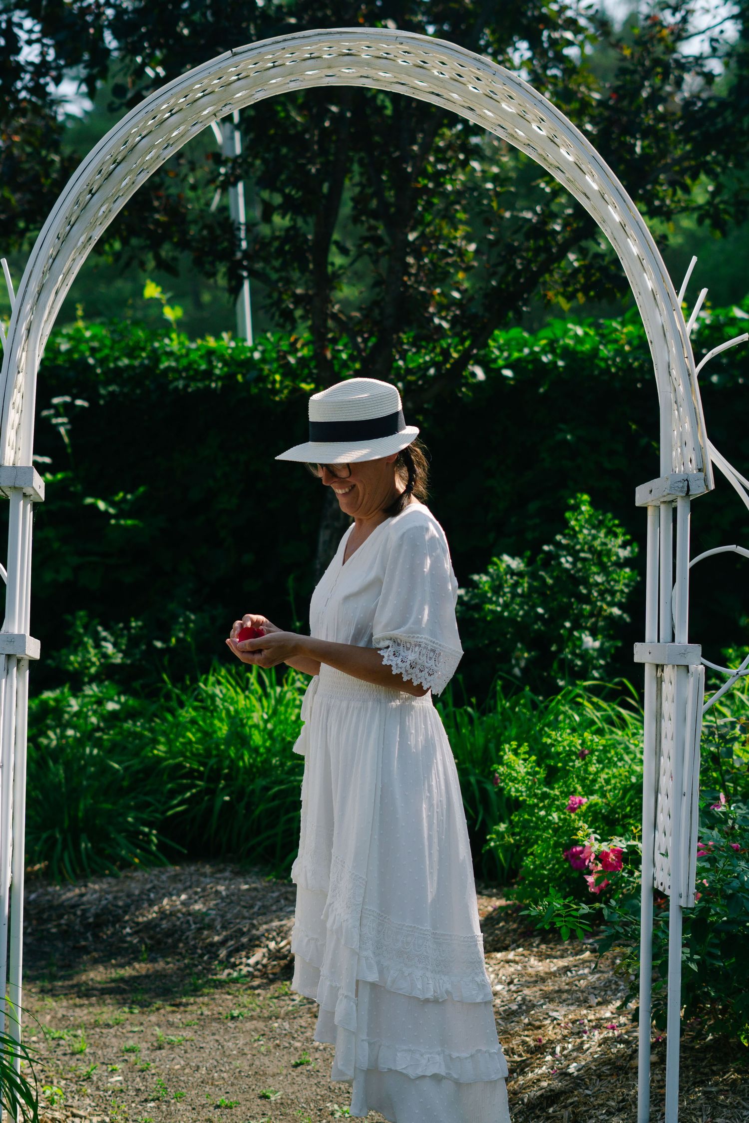 A series of white garden arches frame someone in a flowing white dress walking through a lush outdoor garden setting.