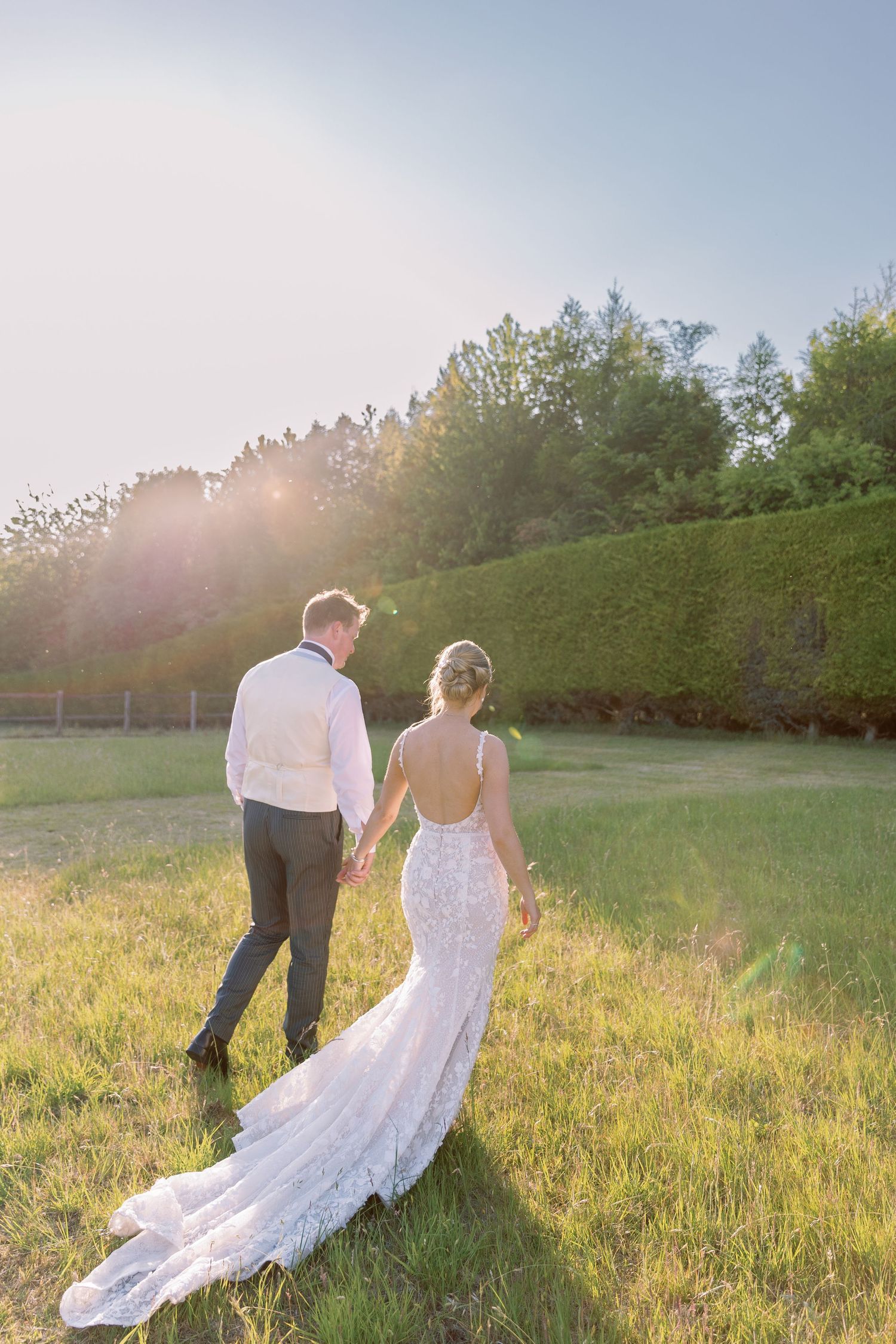 A couple in wedding attire walks through a sunlit meadow while the bride's white lace dress trails behind them.