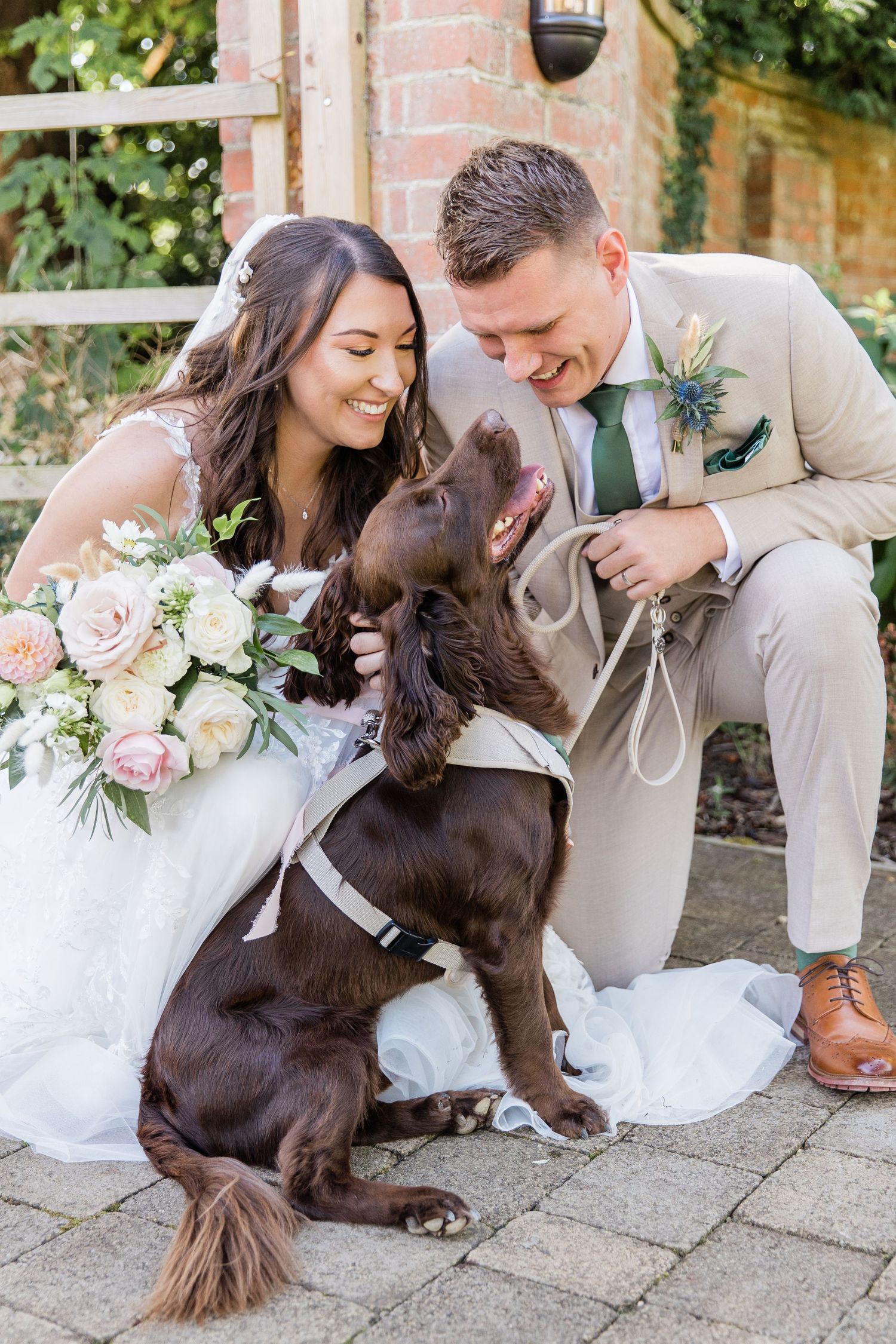 Joyful couple shares tender moment with their brown dog wearing a bow tie during outdoor wedding photo session.