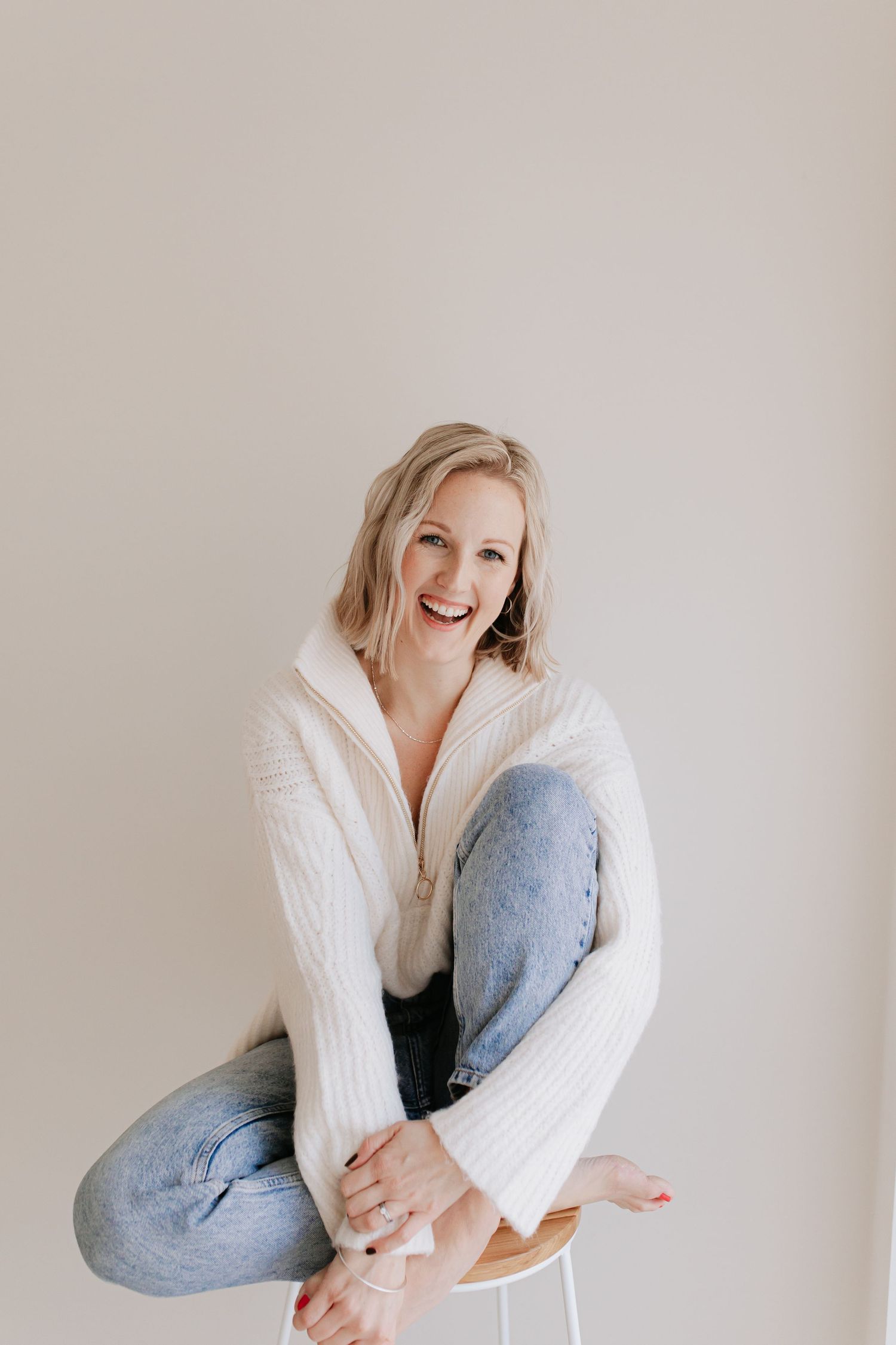 Hampshire Photographer Maddie wearing a white sweater and jeans sits casually against a neutral wall while laughing and smiling.