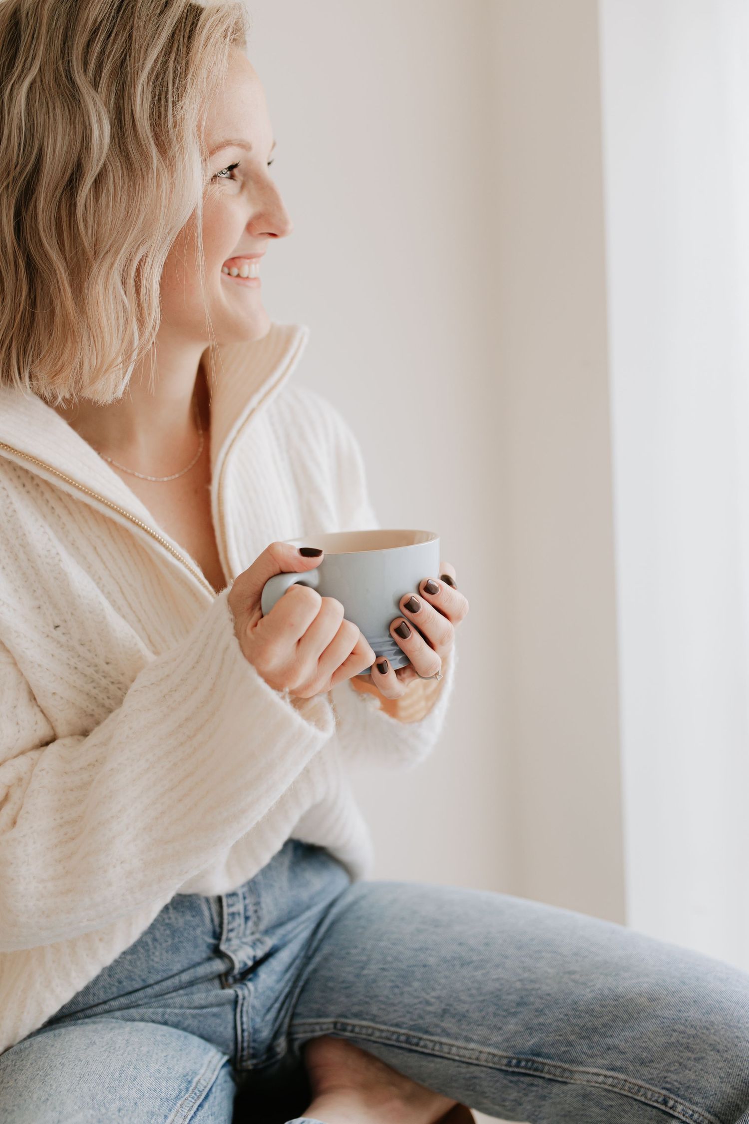 Hampshire Photographer Maddie in cozy white sweater and jeans sips from mug while relaxing at home in natural light.