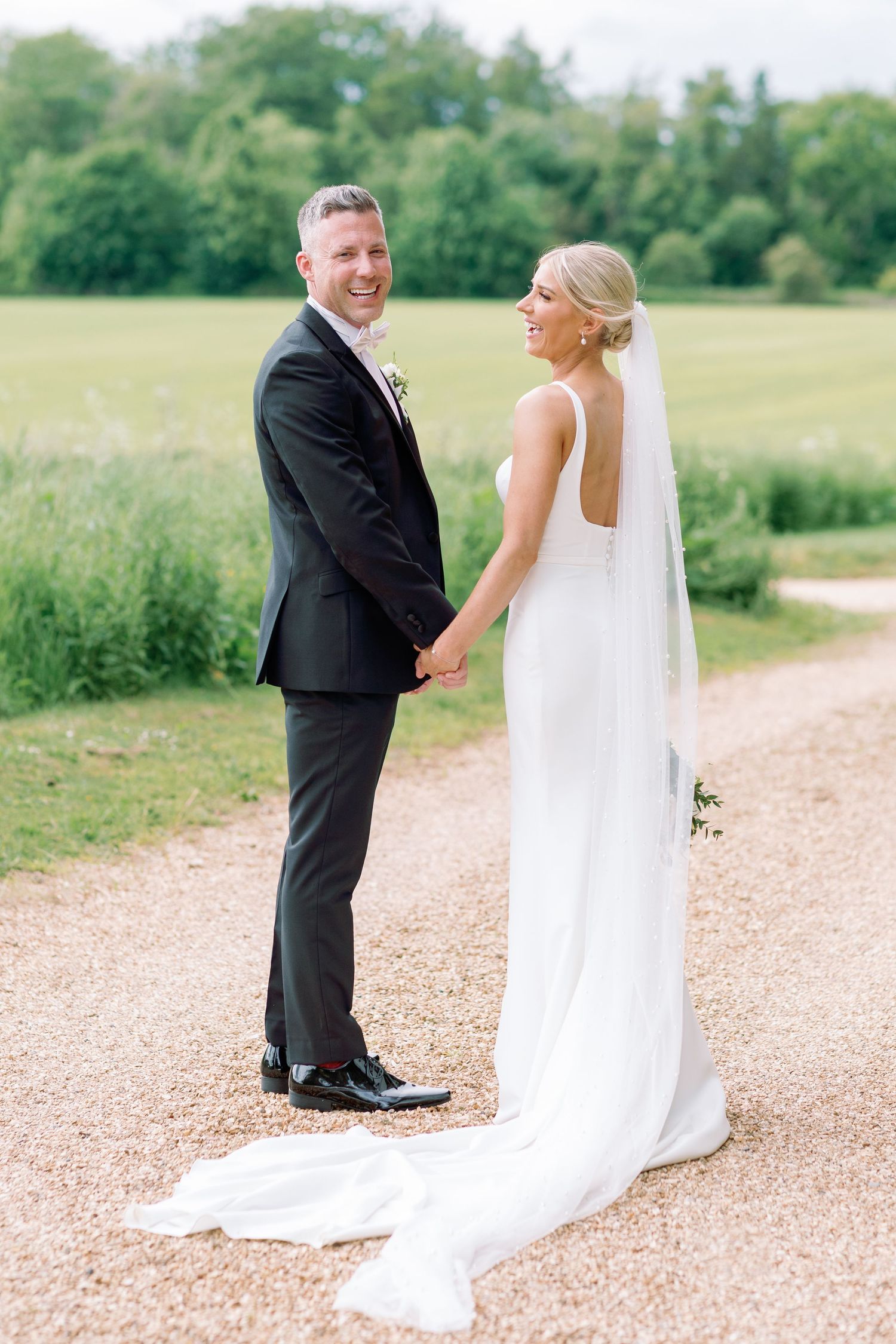 A couple in wedding attire stands on a gravel path surrounded by green fields on their special day.