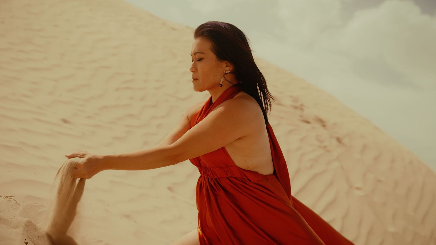 Woman in red halter dress poses gracefully on golden sand dunes in a serene desert landscape.