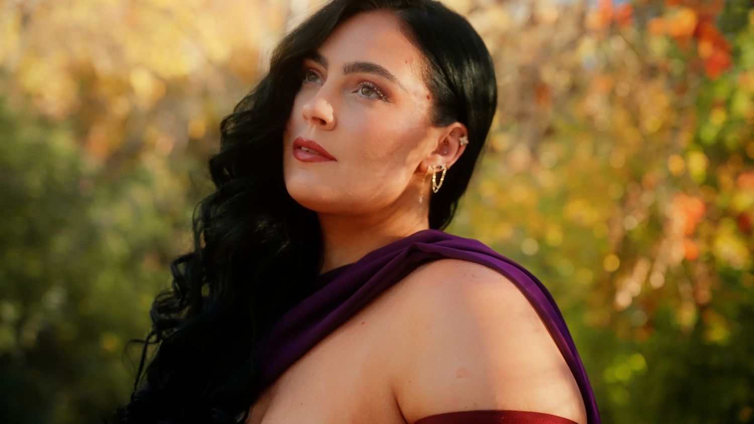 A person in a red dress poses outdoors against a backdrop of autumn foliage during golden hour.