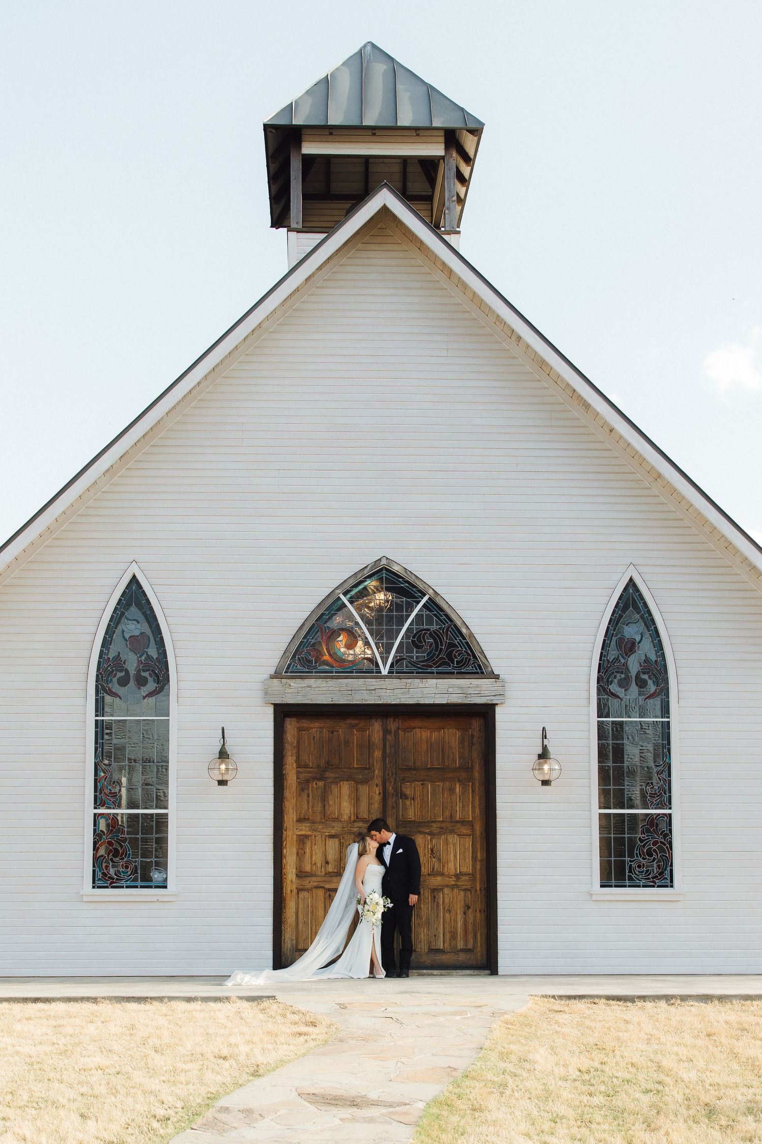 A small white chapel with stained glass windows and wooden doors stands against a bright sky.