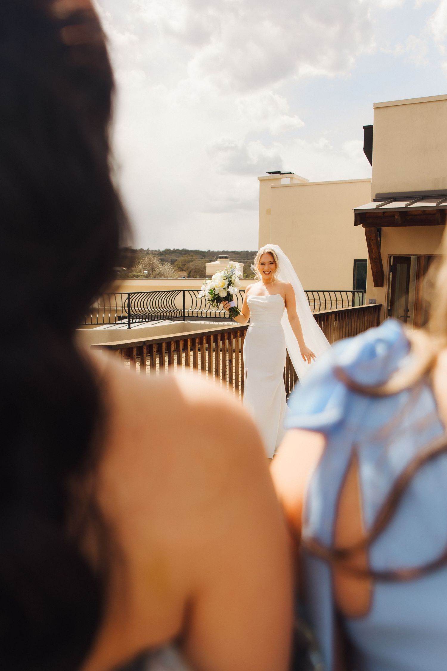 A bride in a white wedding dress stands on a rooftop terrace during a sunny day.