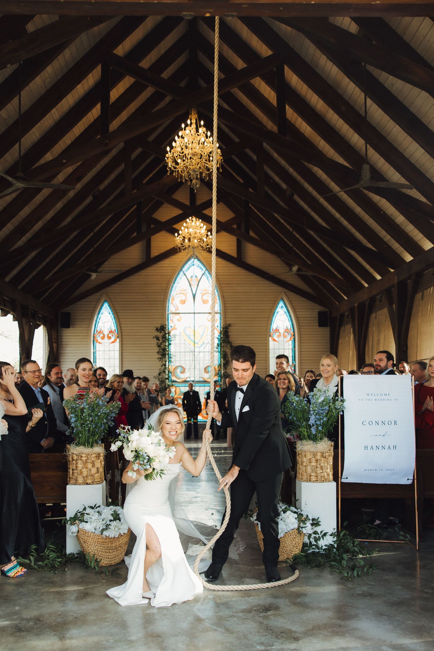 Wedding couple celebrating in front of decorative floral arrangements in a chapel with exposed wooden beams.