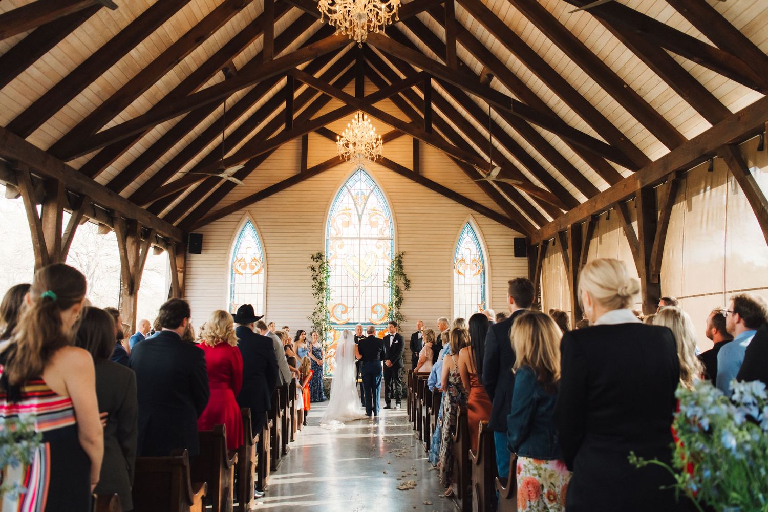 A wedding ceremony in a rustic wooden church with exposed beams and stained glass windows illuminated by natural light.