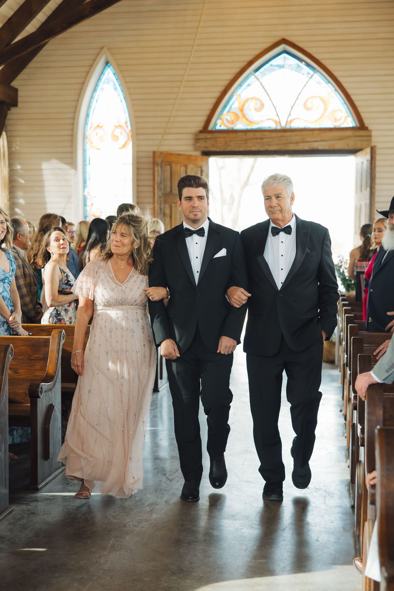 Formal wedding processional inside a historic chapel with arched windows and wooden pews.