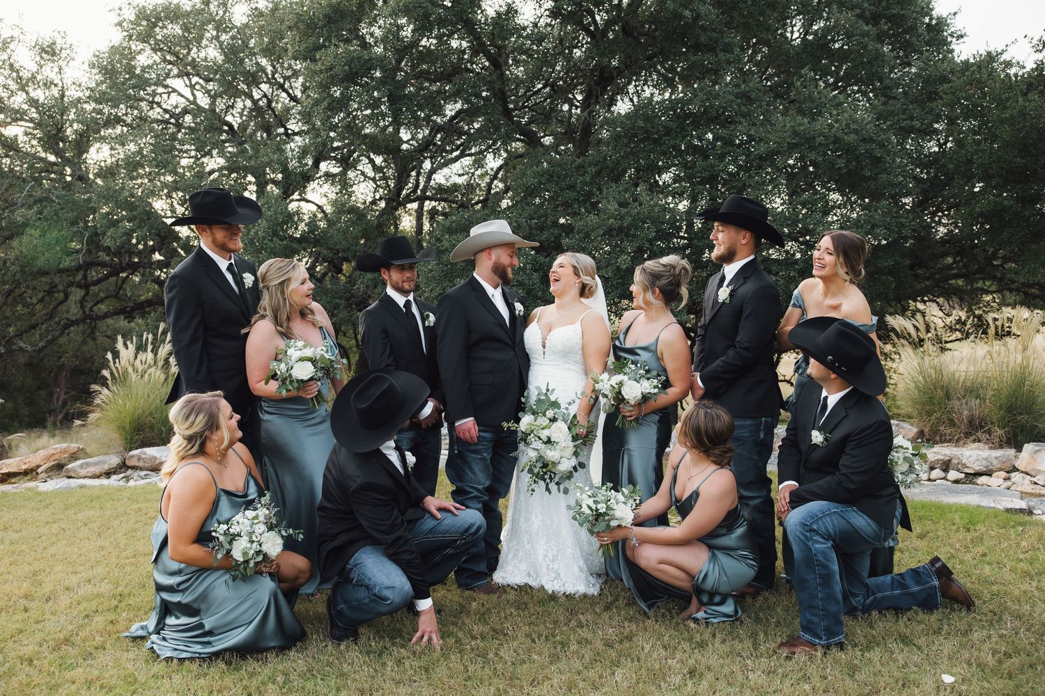 Wedding party poses casually on grassy lawn with trees in background, wearing formal attire and denim.