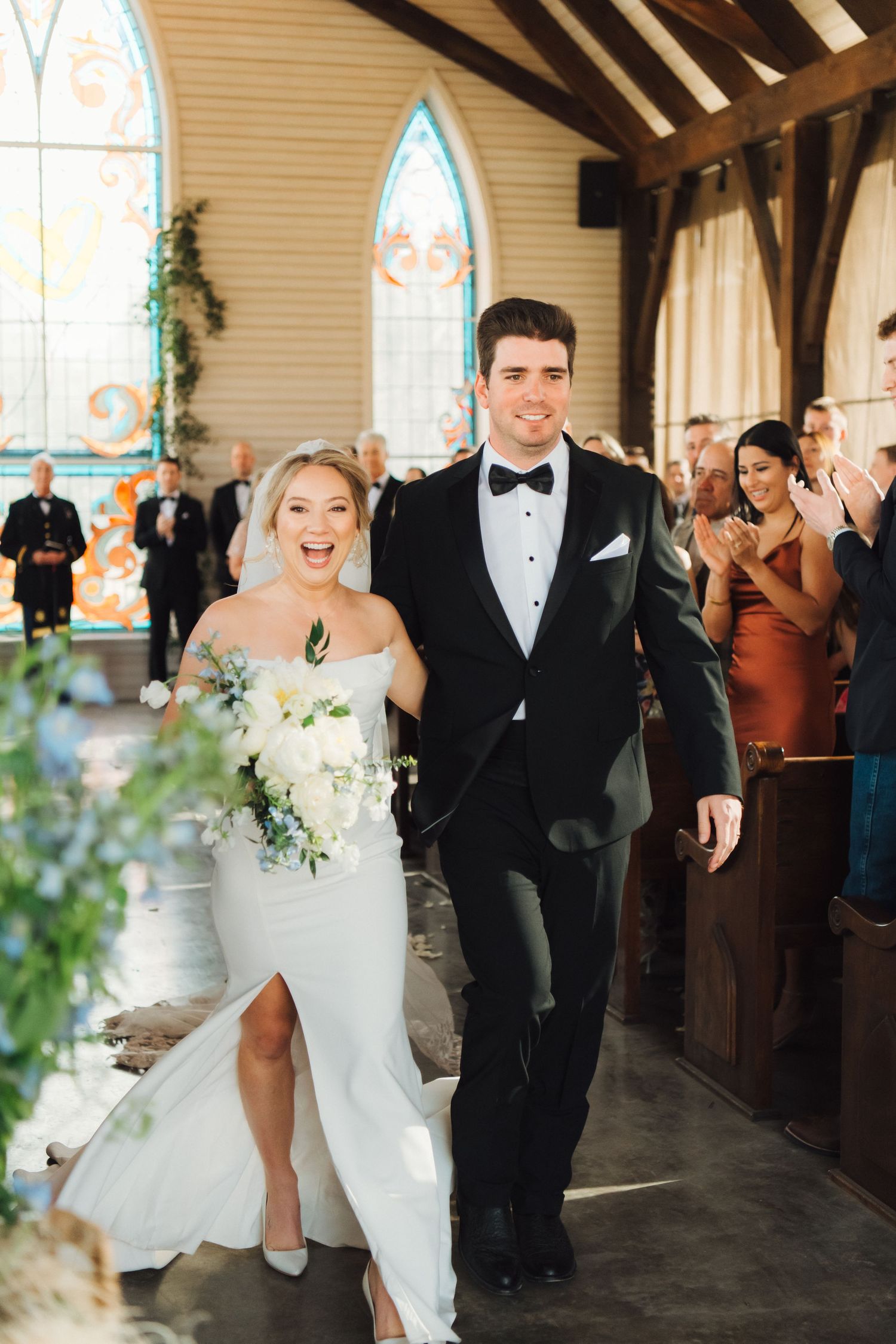 Wedding party celebrates joyfully while walking down aisle at church ceremony with stained glass windows and floral decor.