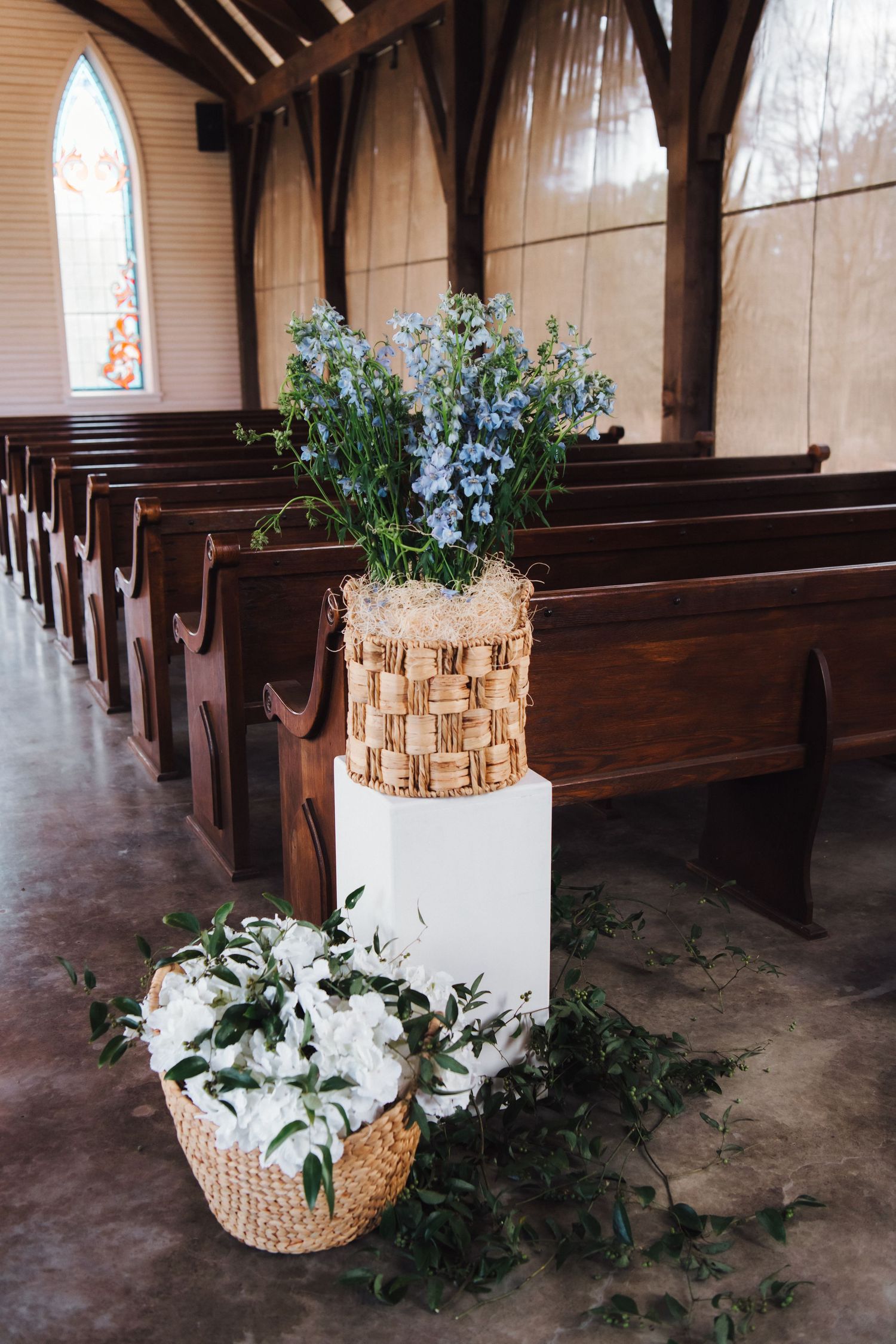 Rustic church aisle decorated with blue flowers in a birch vase and white floral arrangement in basket near wooden pews.