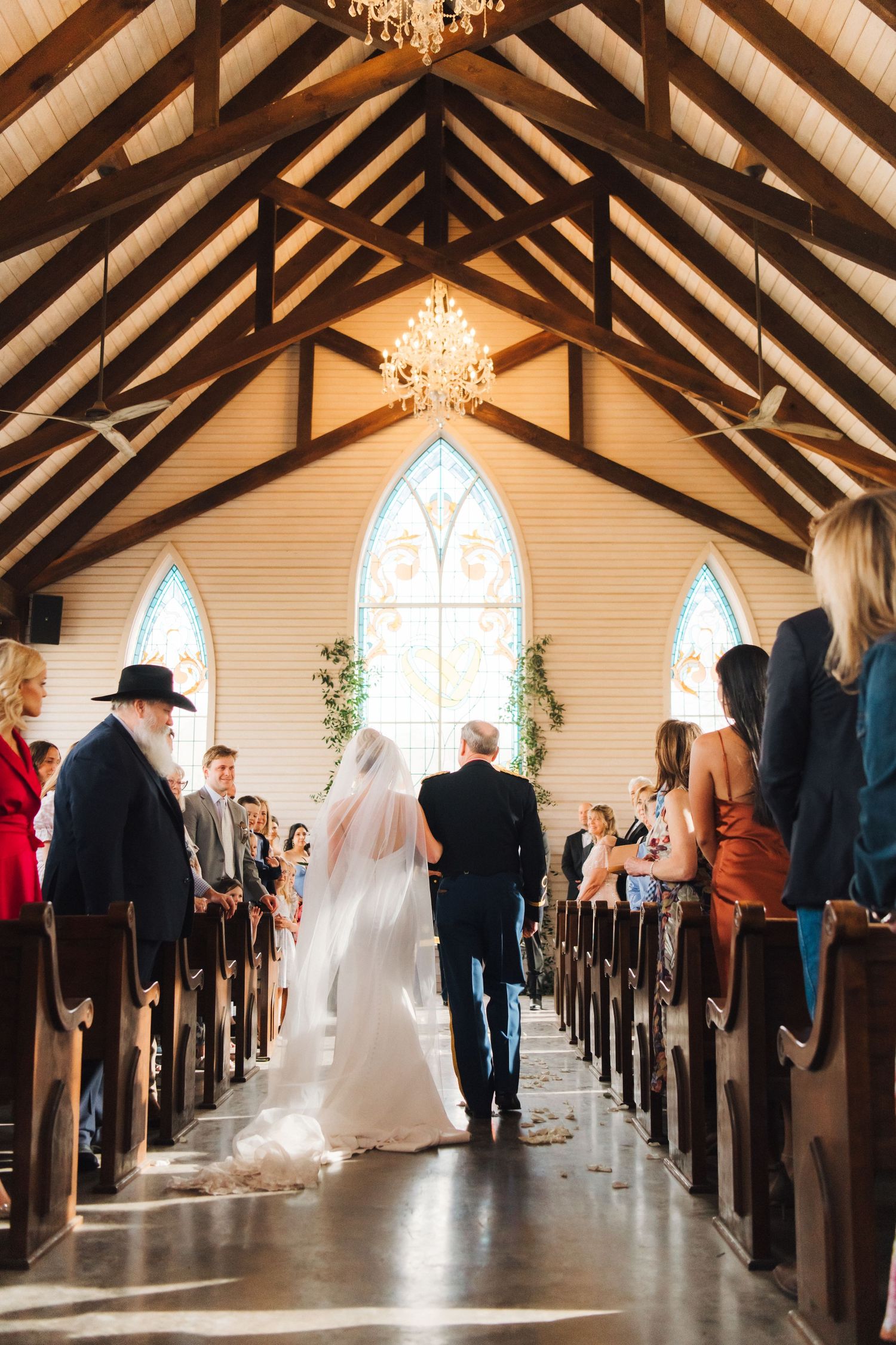 A bride in white gown walks down the aisle of a rustic chapel with vaulted wooden beams and stained glass windows.
