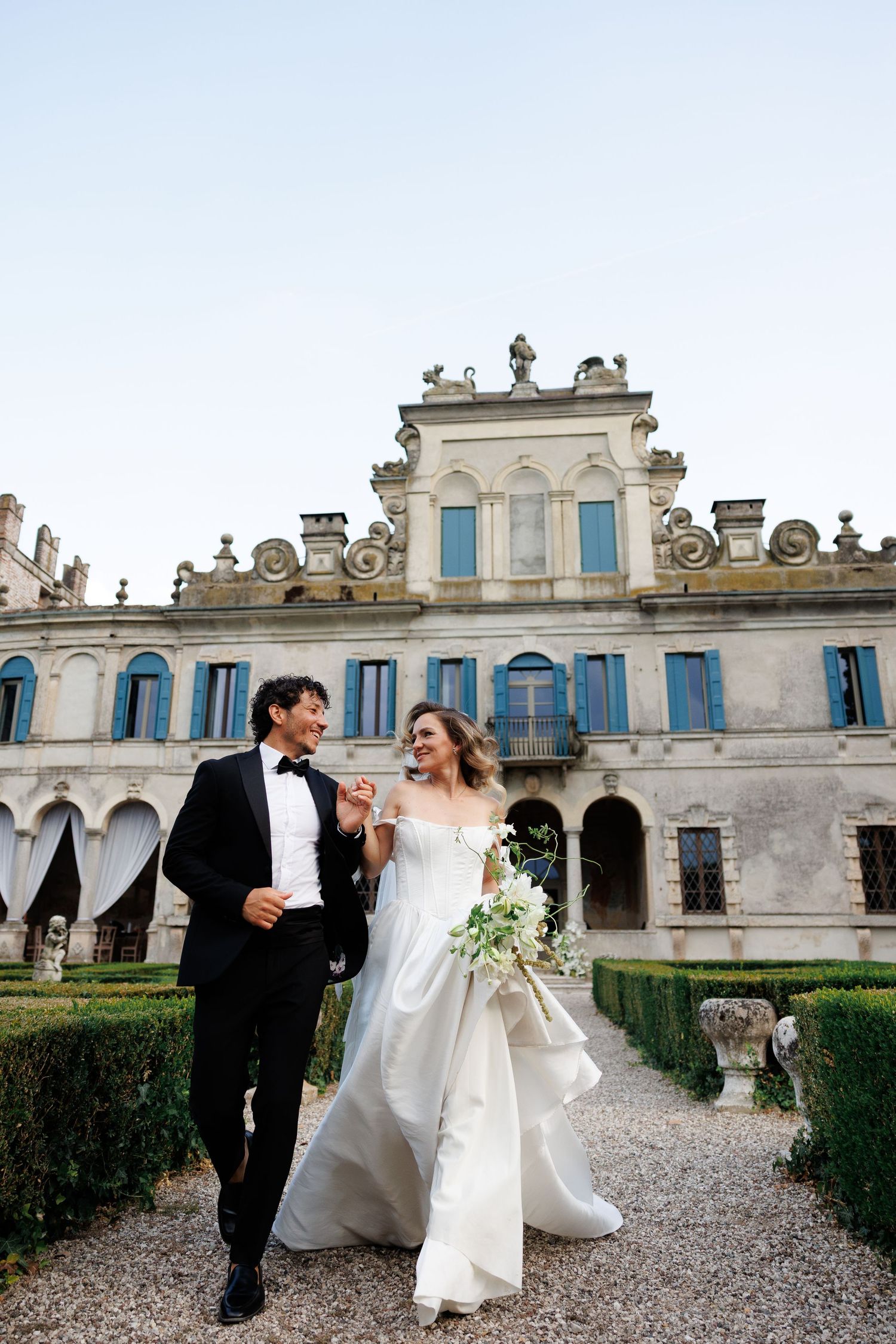 A couple in formal wedding attire share romantic moments on a gravel path in front of a grand historic mansion.