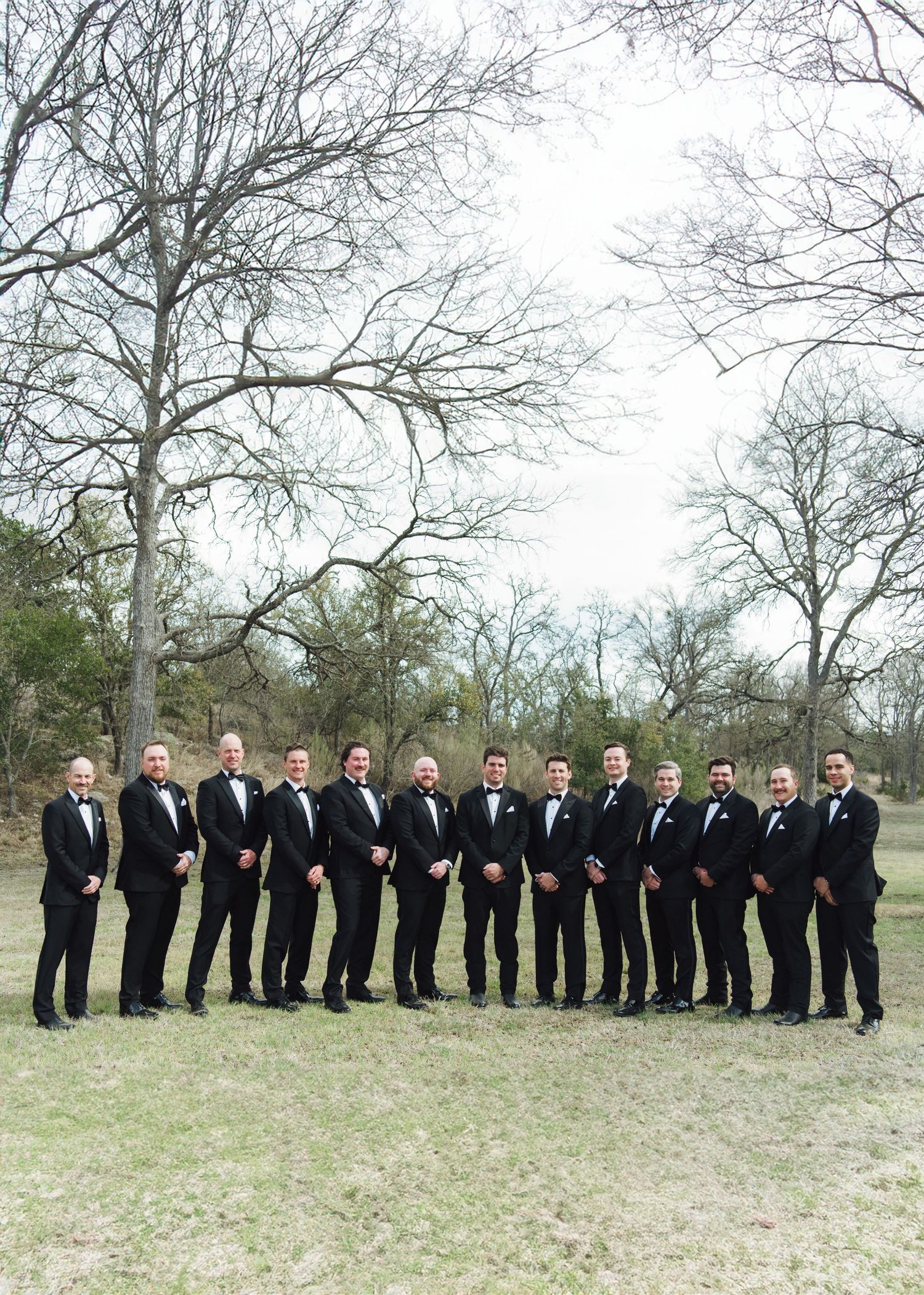 A group of groomsmen in black tuxedos pose outdoors under bare winter trees on a cloudy day.