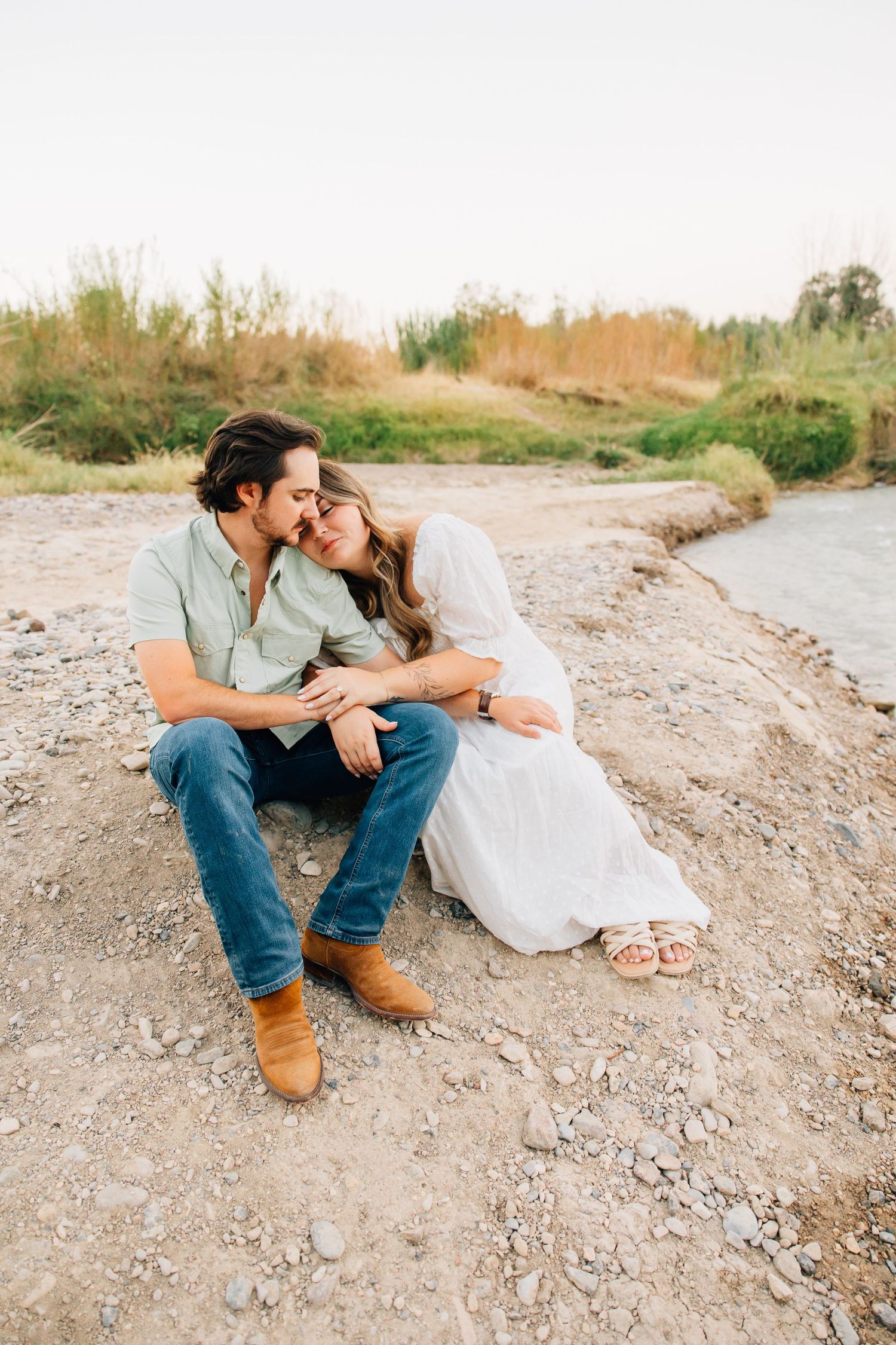 A couple in casual attire sits intimately together on a rocky riverbank during a romantic photoshoot.