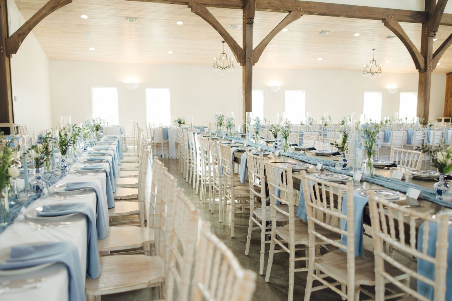 A wedding reception venue with white chairs and light blue tablecloths set in a rustic barn-style space with wooden beams.