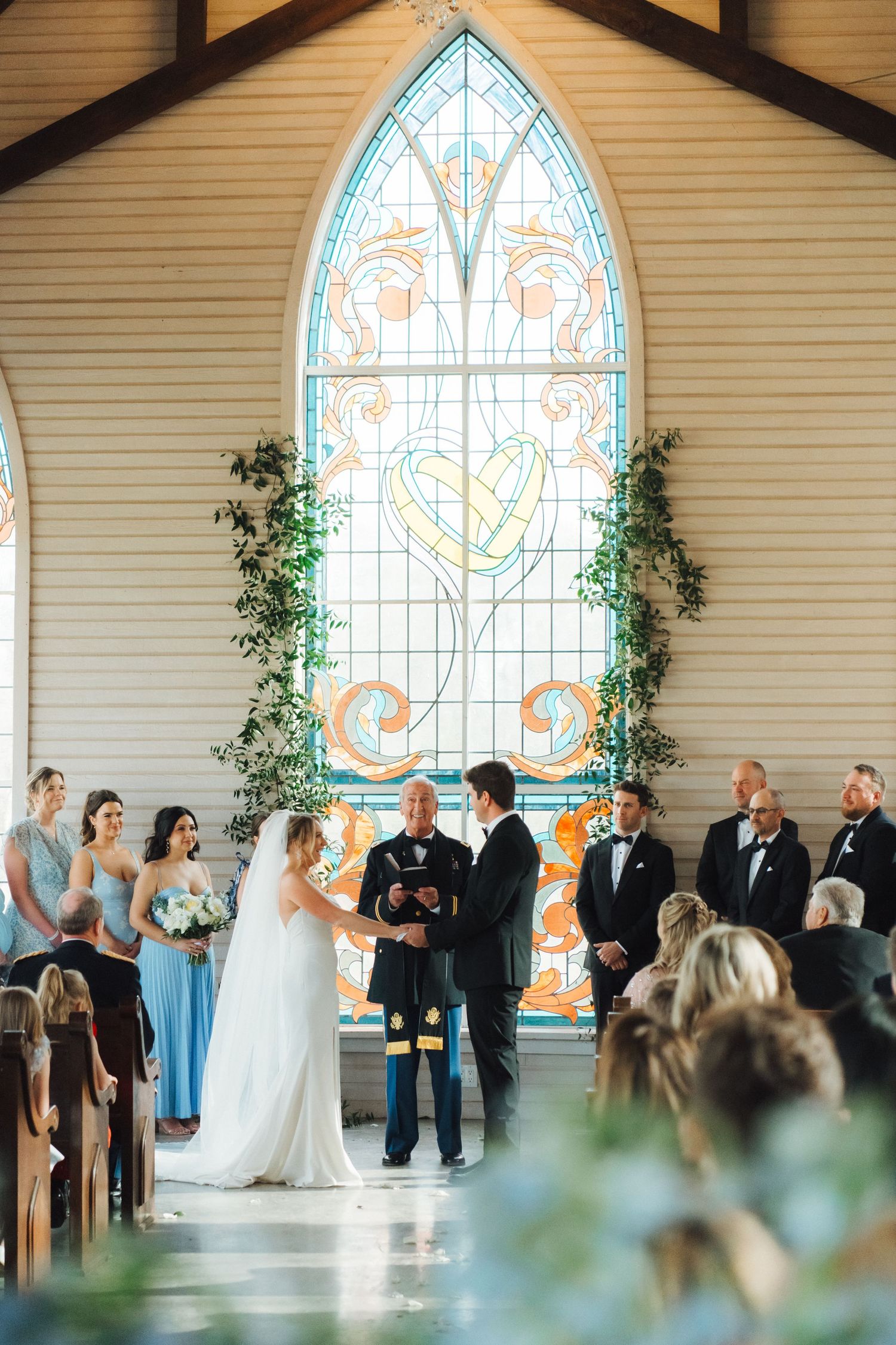 A wedding ceremony in progress inside a church with beautiful stained glass windows and greenery decor.
