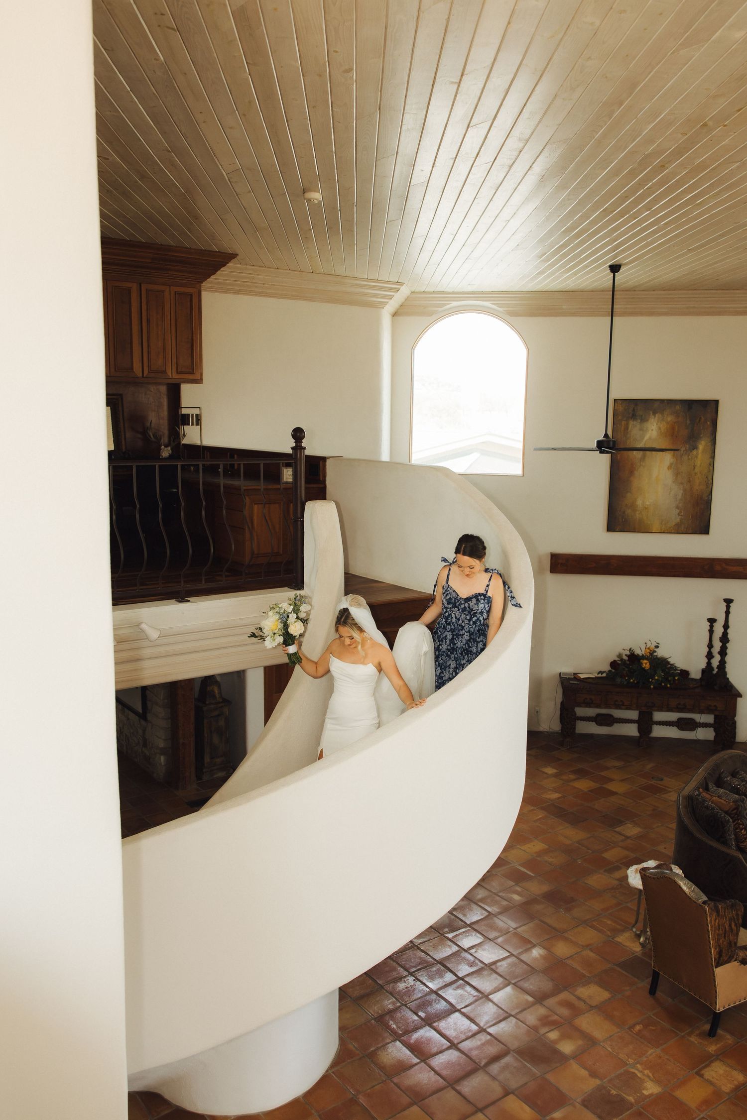 People seated on a curved white staircase with wooden ceiling and terracotta tile floor in an indoor space.