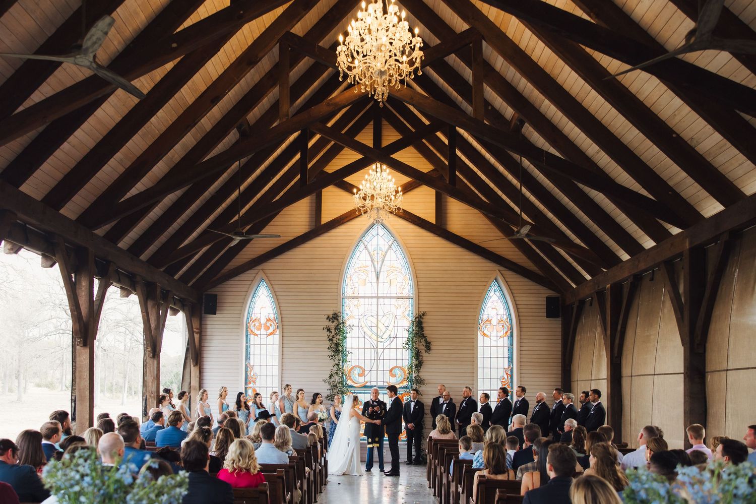 A grand chapel with exposed wooden beams, gothic windows and a crystal chandelier hosts a wedding ceremony gathering.