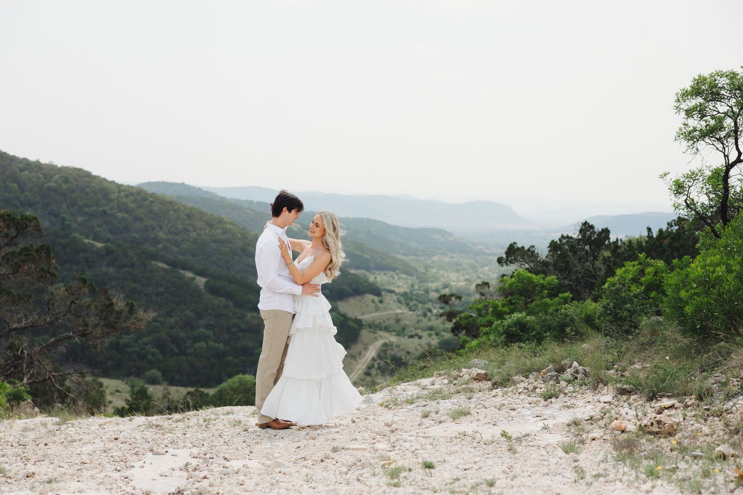 Bride and groom share an intimate moment while sitting on a rocky cliff overlooking mountain landscapes at sunset.