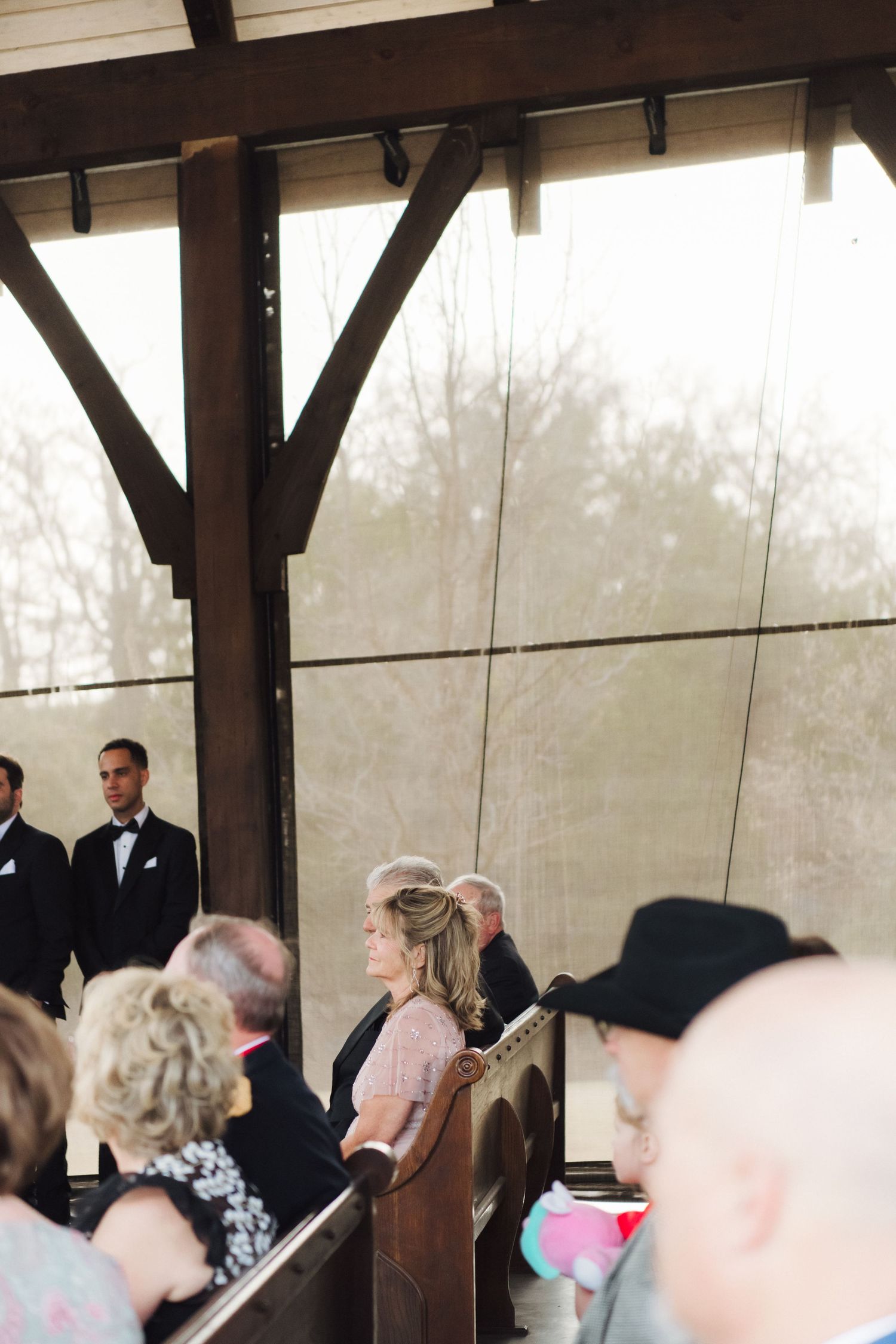 Wedding guests gather under a wooden pavilion during an outdoor ceremony.