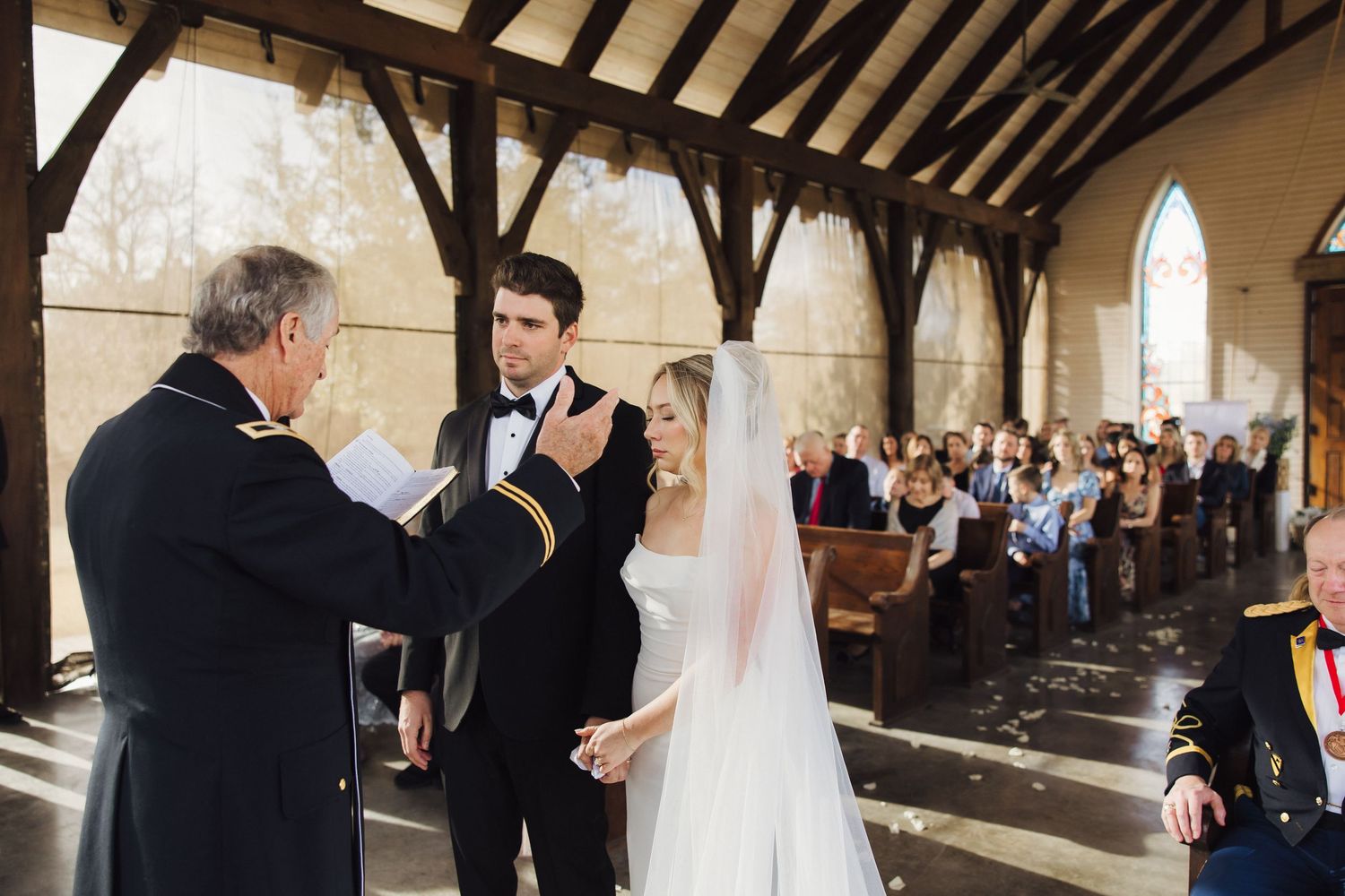 A wedding ceremony takes place in a covered outdoor venue with exposed wooden beams and stone archways.