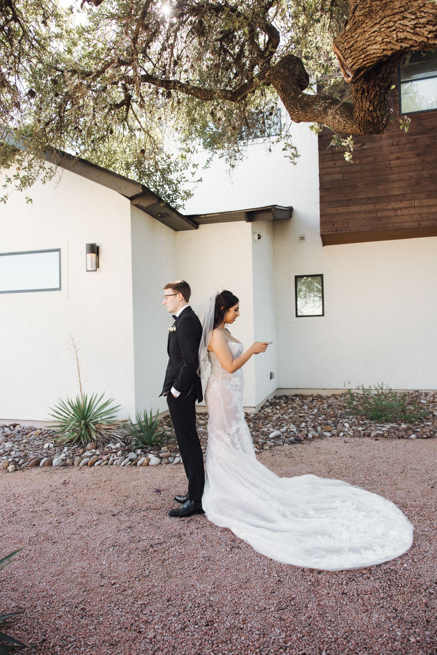 A bride in a white lace gown with long train stands back-to-back with groom in black suit during first look moment.