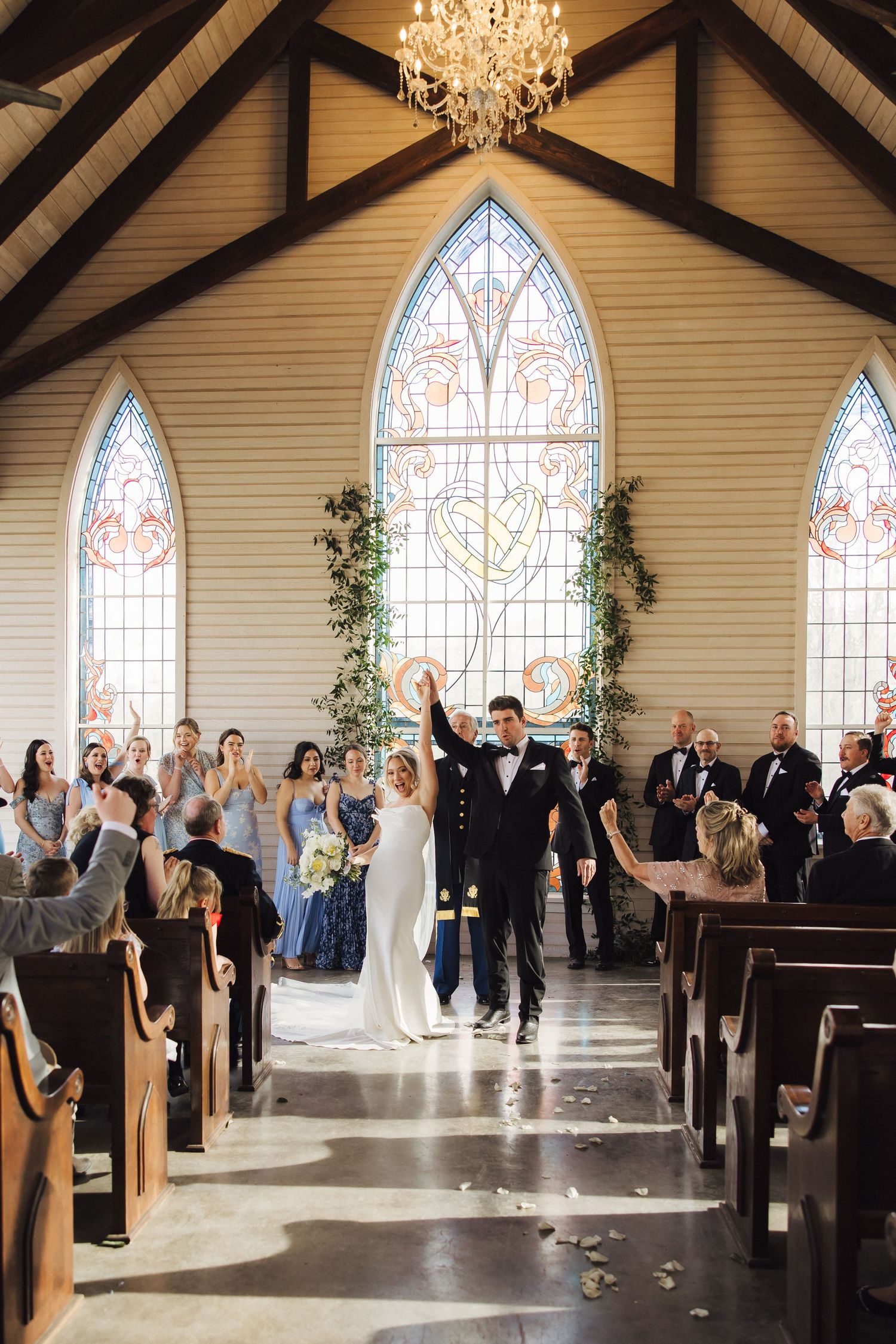 Newlyweds celebrate with raised hands at the altar while guests applaud in a beautiful chapel setting.