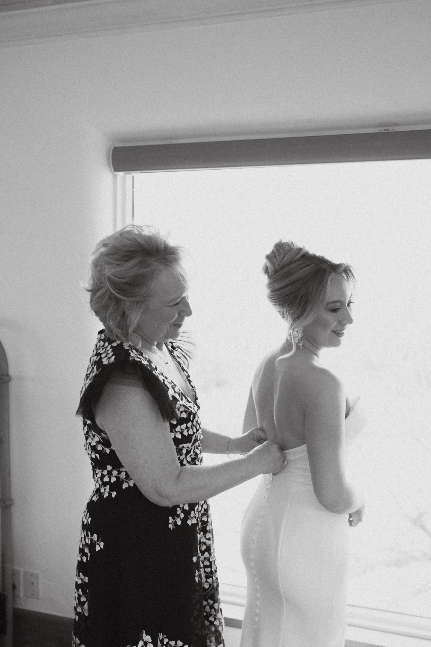 Black and white photograph capturing a tender moment during wedding dress preparations.