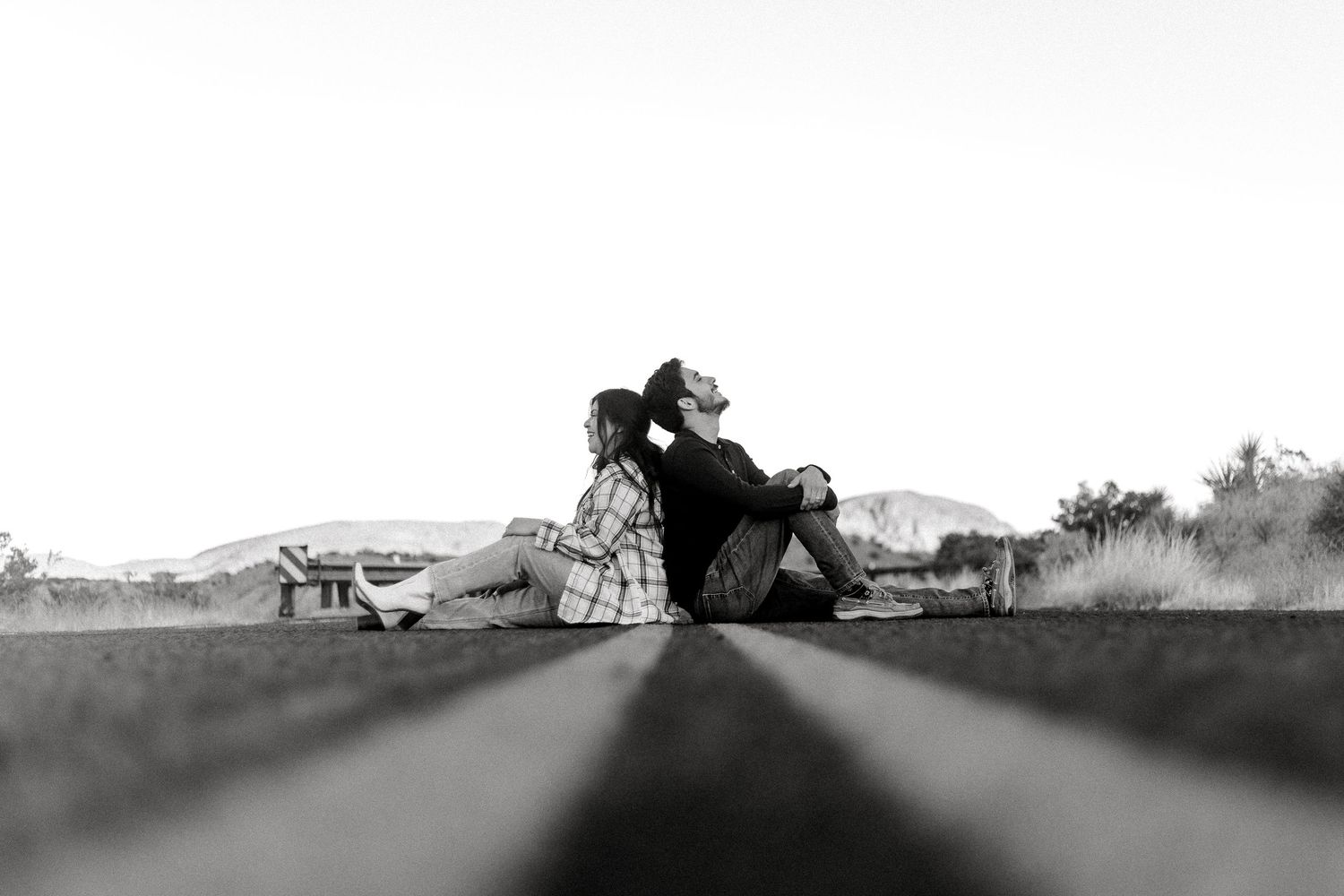 A black and white photo of a couple sitting close together on a road, shot from a low angle perspective.