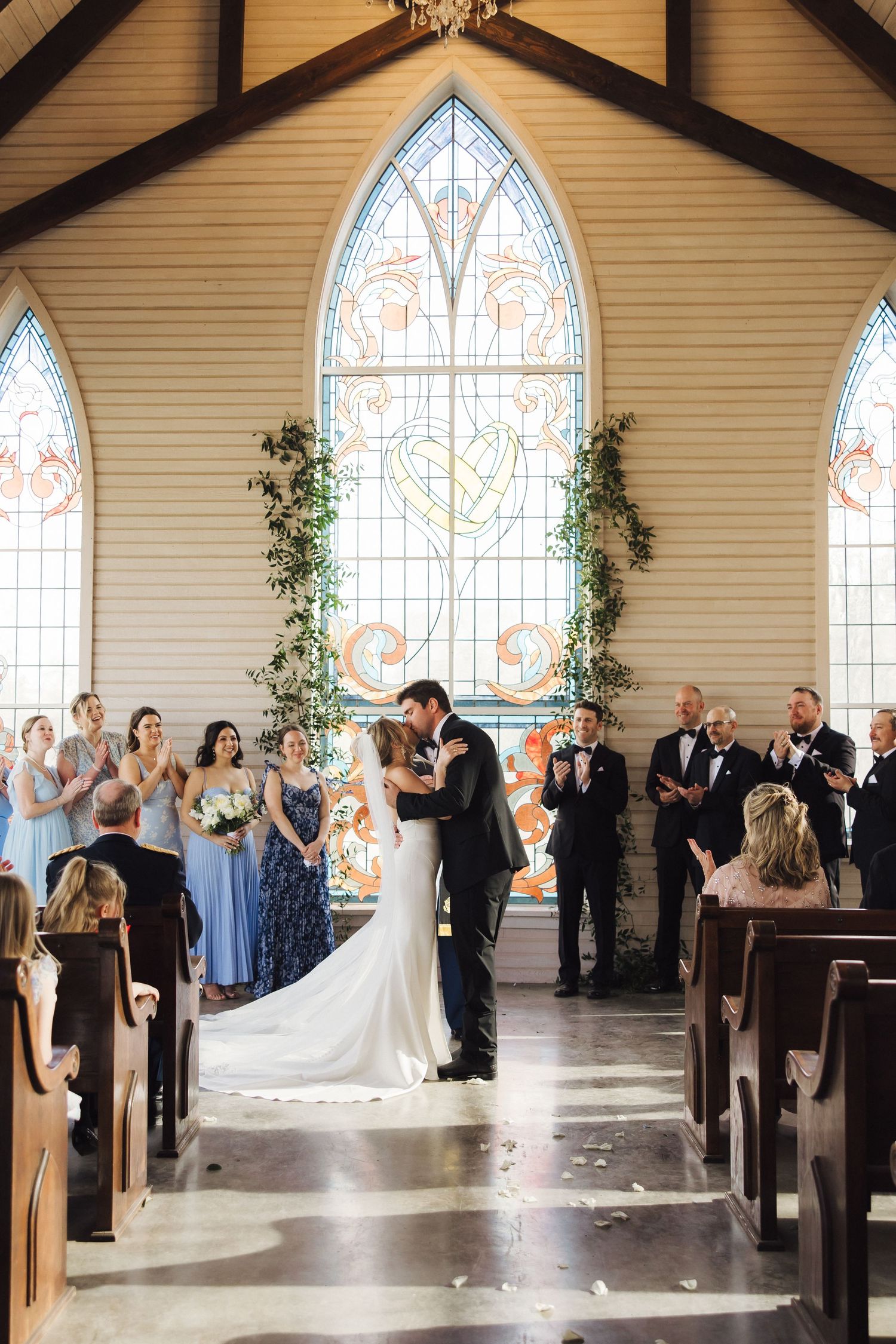 A wedding ceremony takes place in a chapel with stained glass windows and greenery-adorned arches.