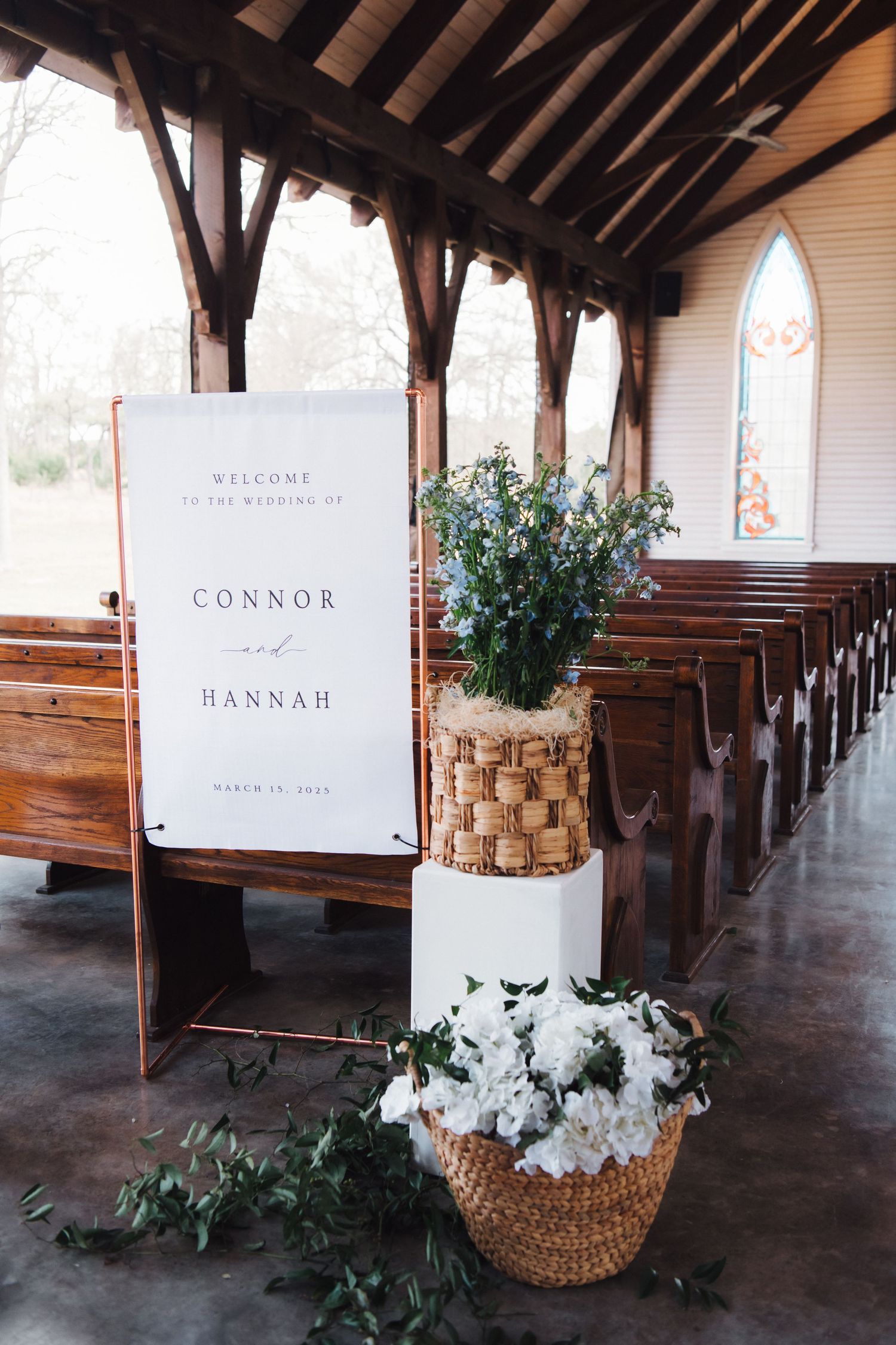 Wedding welcome sign for Connor and Hannah displayed in rustic chapel with floral arrangements.