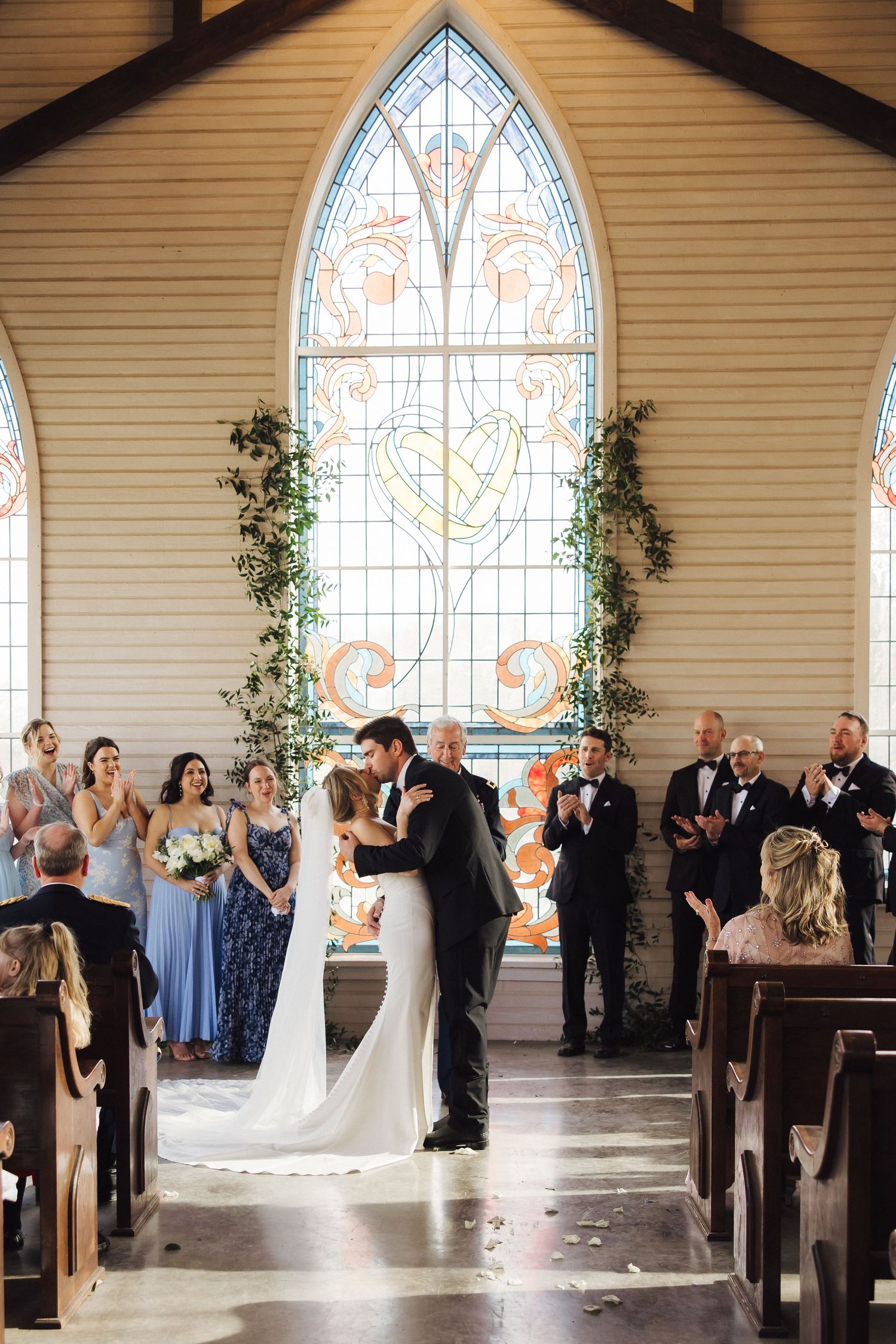 Wedding ceremony in progress inside a chapel with elegant arched stained glass windows and wooden pews.