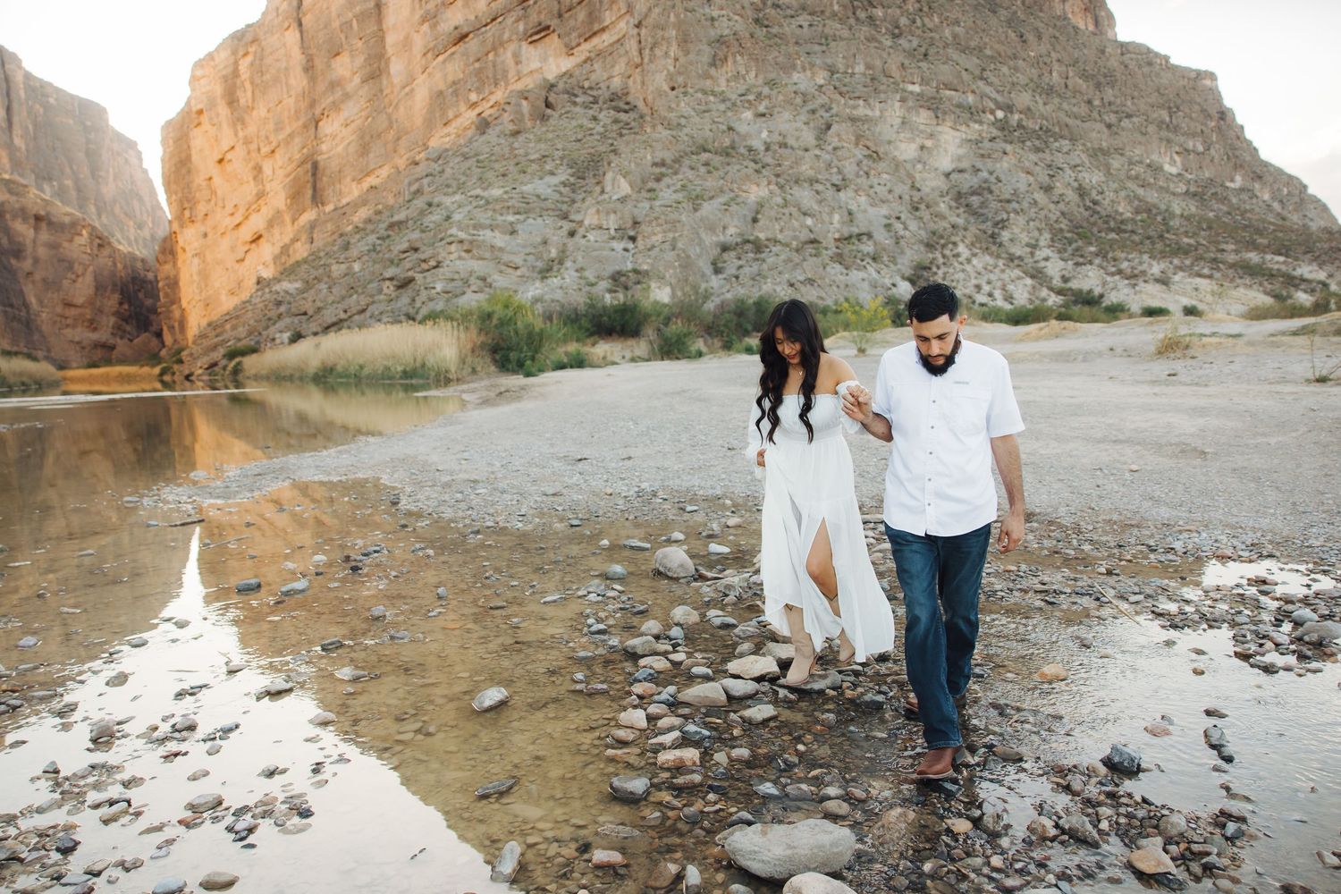 A couple walks together along a rocky shoreline beneath towering canyon walls in a scenic desert landscape.